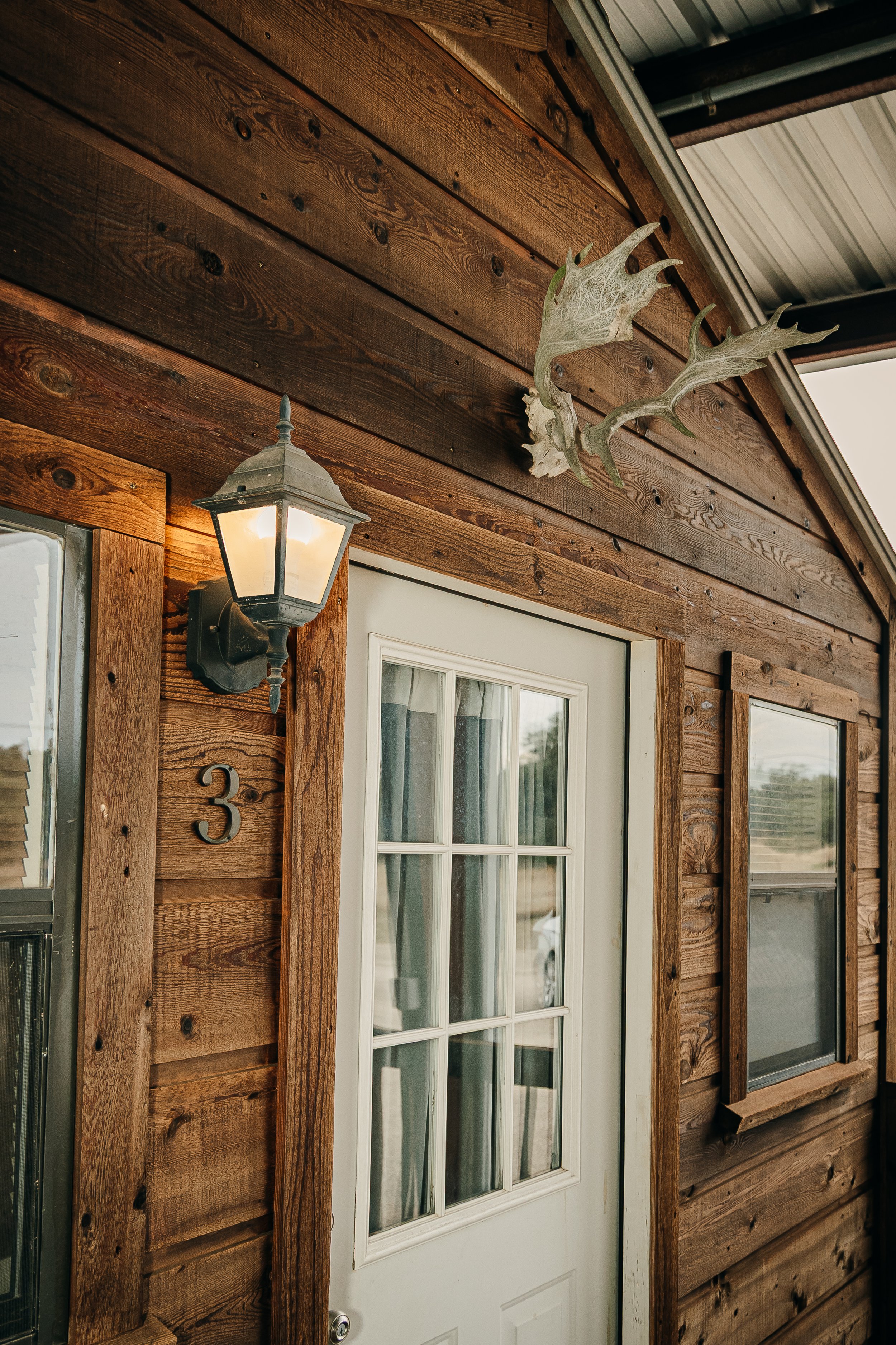 Wooden house exterior with a white door, a lantern light, house number 3, and a mounted deer antler decor piece above the door.