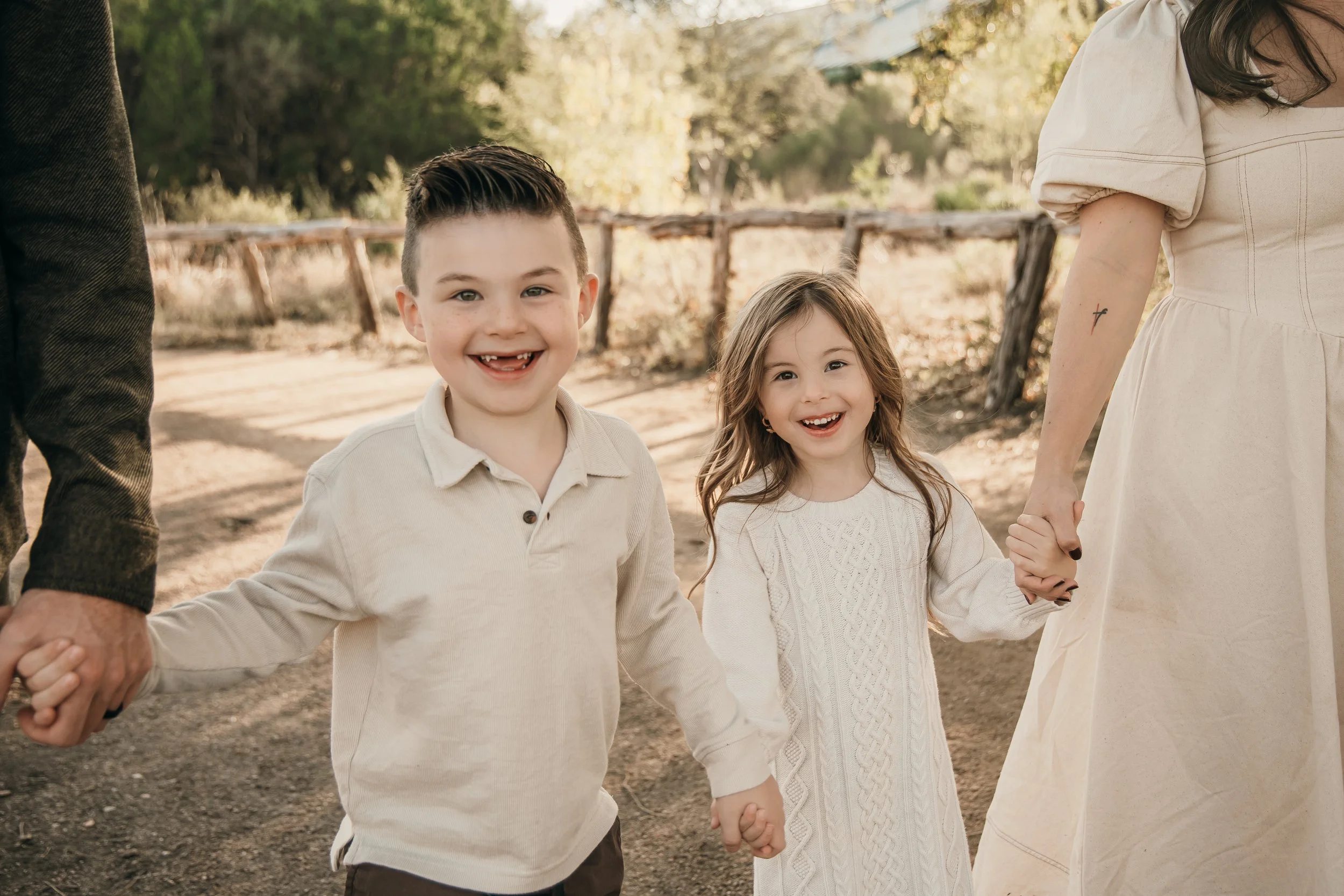 Two young children, a boy and a girl, holding hands with adults outdoors on a dirt path surrounded by trees.