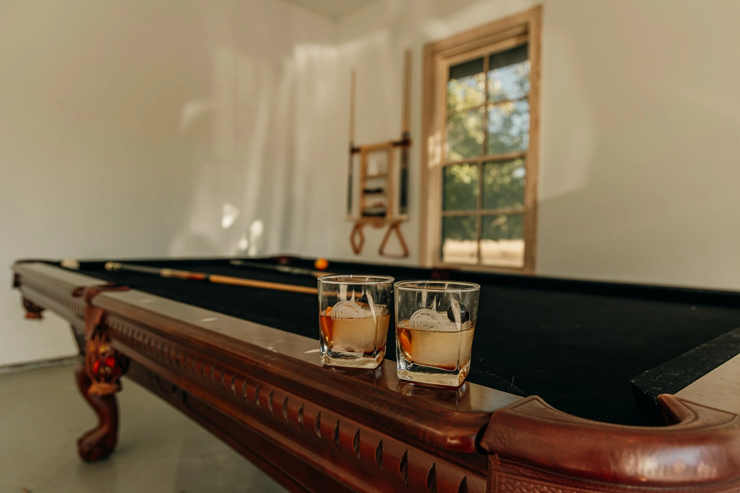 Two glasses with whiskey and ice on a pool table's edge, in a room with a window showing greenery outside.