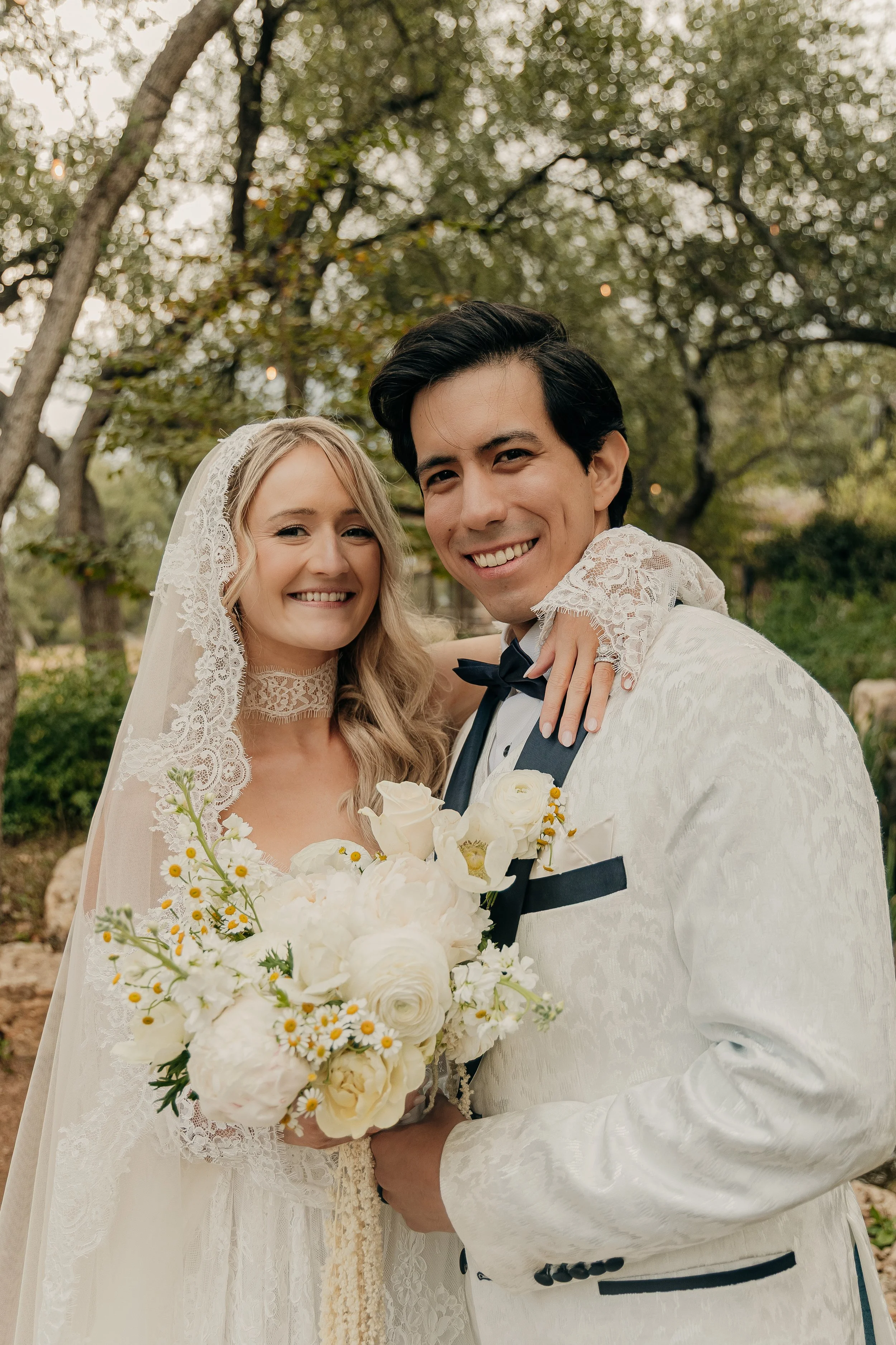 A happy couple in wedding attire posing outdoors with trees and greenery in the background. The bride holds a bouquet of white flowers, and they are smiling at the camera.