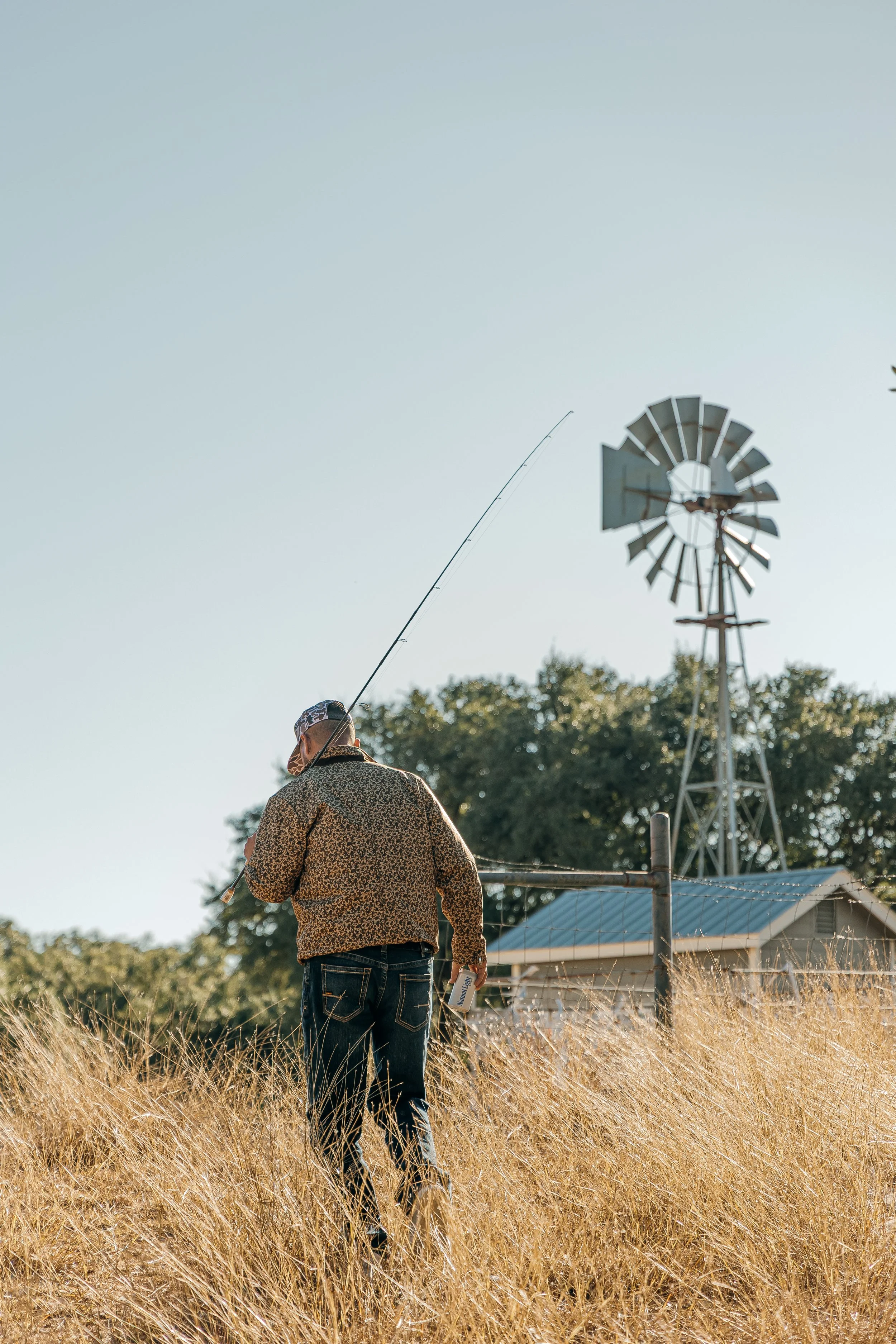 A person walking through a field of tall, dry grass while holding a fishing rod, with a windmill and a small building in the background under a clear sky.