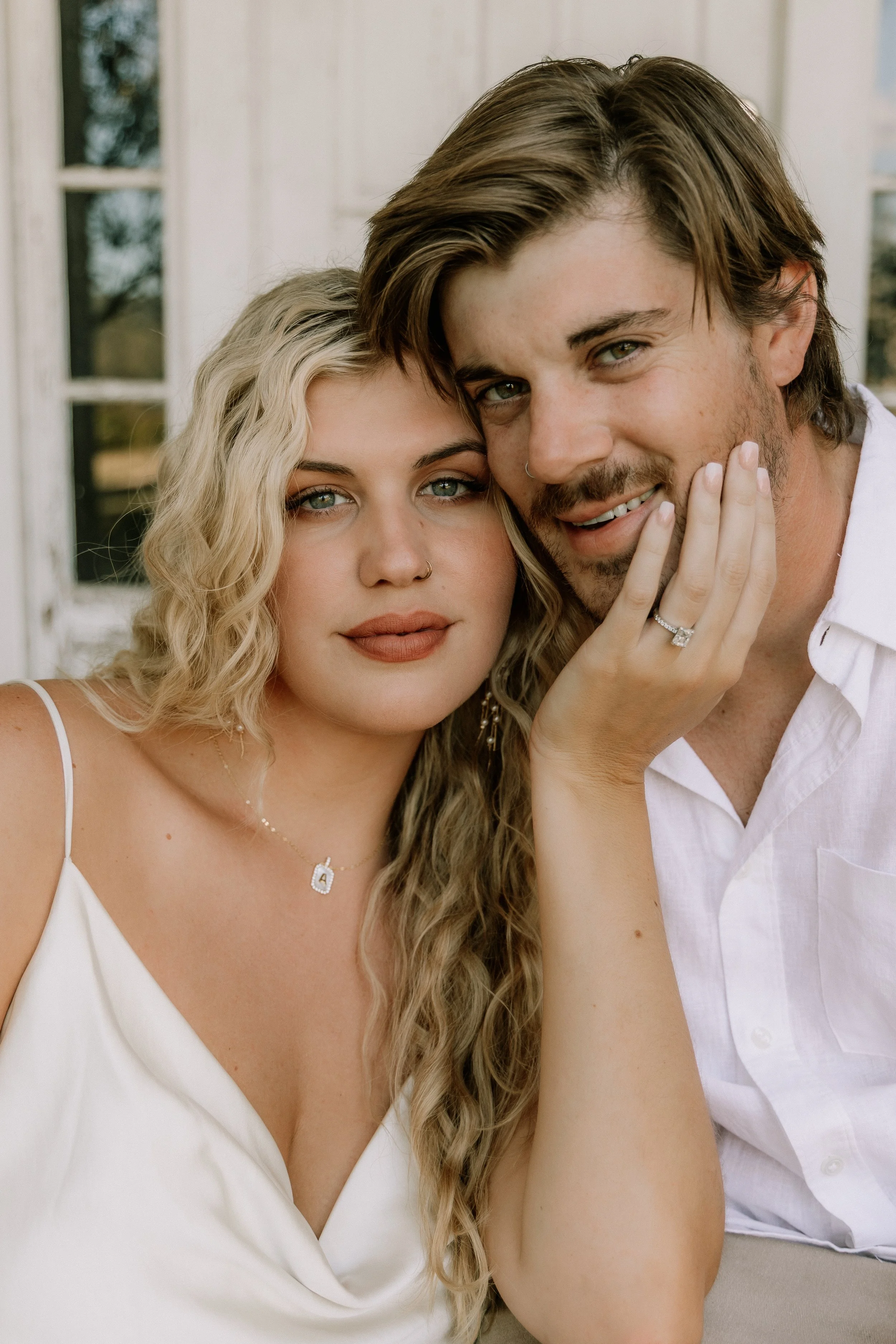 A couple posing closely together, with the woman touching the man's face, both smiling softly, in front of a rustic background.