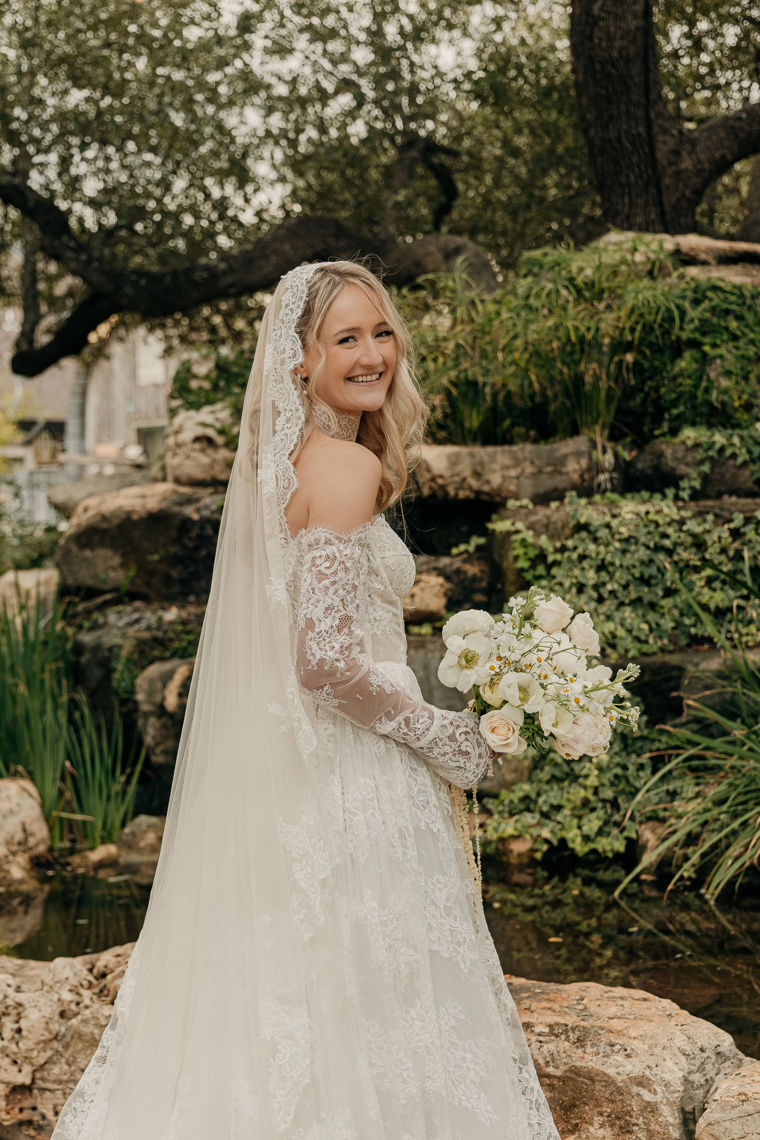 A bride in a lace wedding gown and veil, smiling and holding a bouquet of white and blush flowers, standing outdoors in a garden with rocks and greenery.