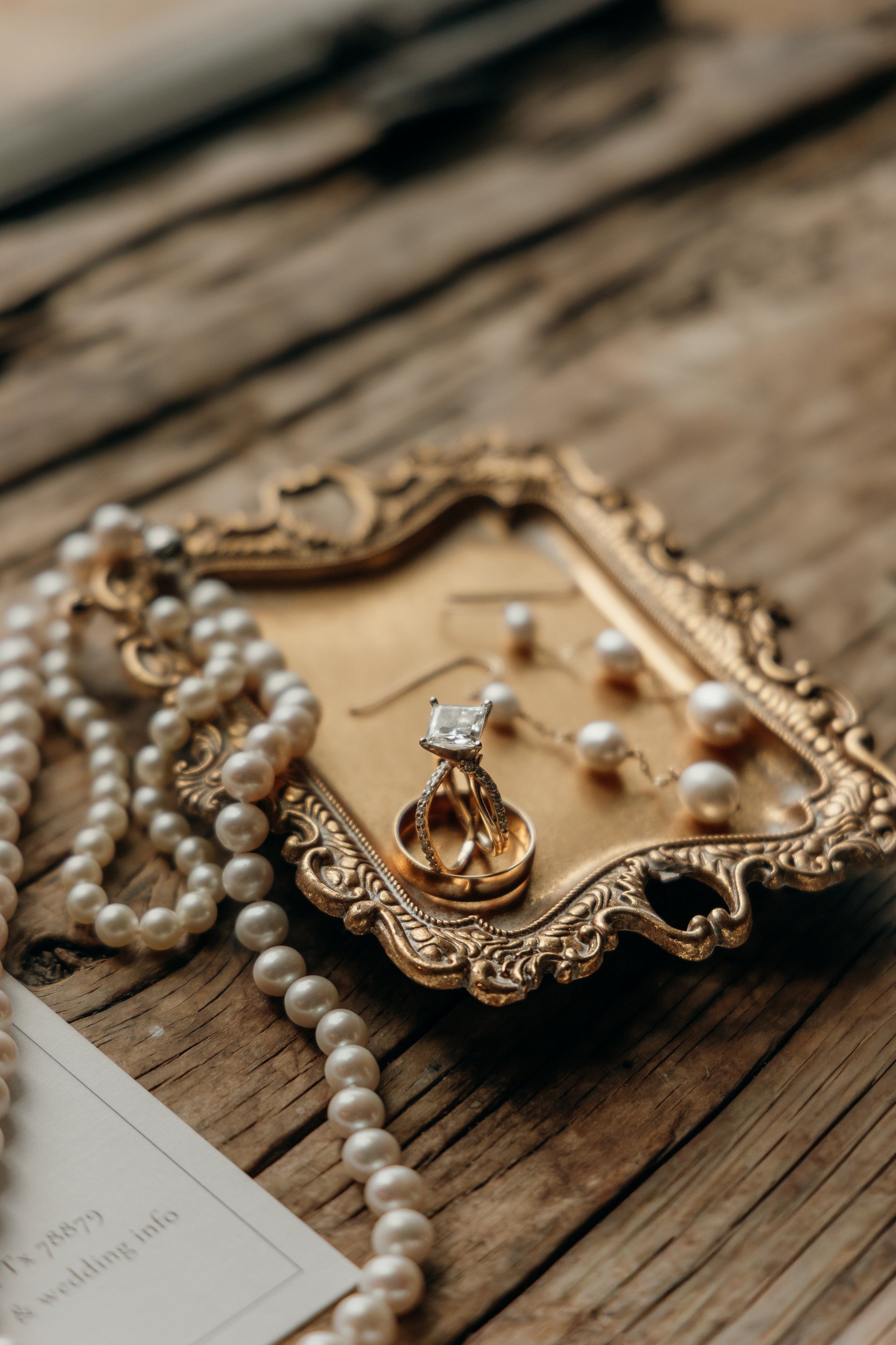 A vintage gold picture frame containing a diamond ring and gold wedding bands, along with pearl jewelry, on a rustic wooden surface.