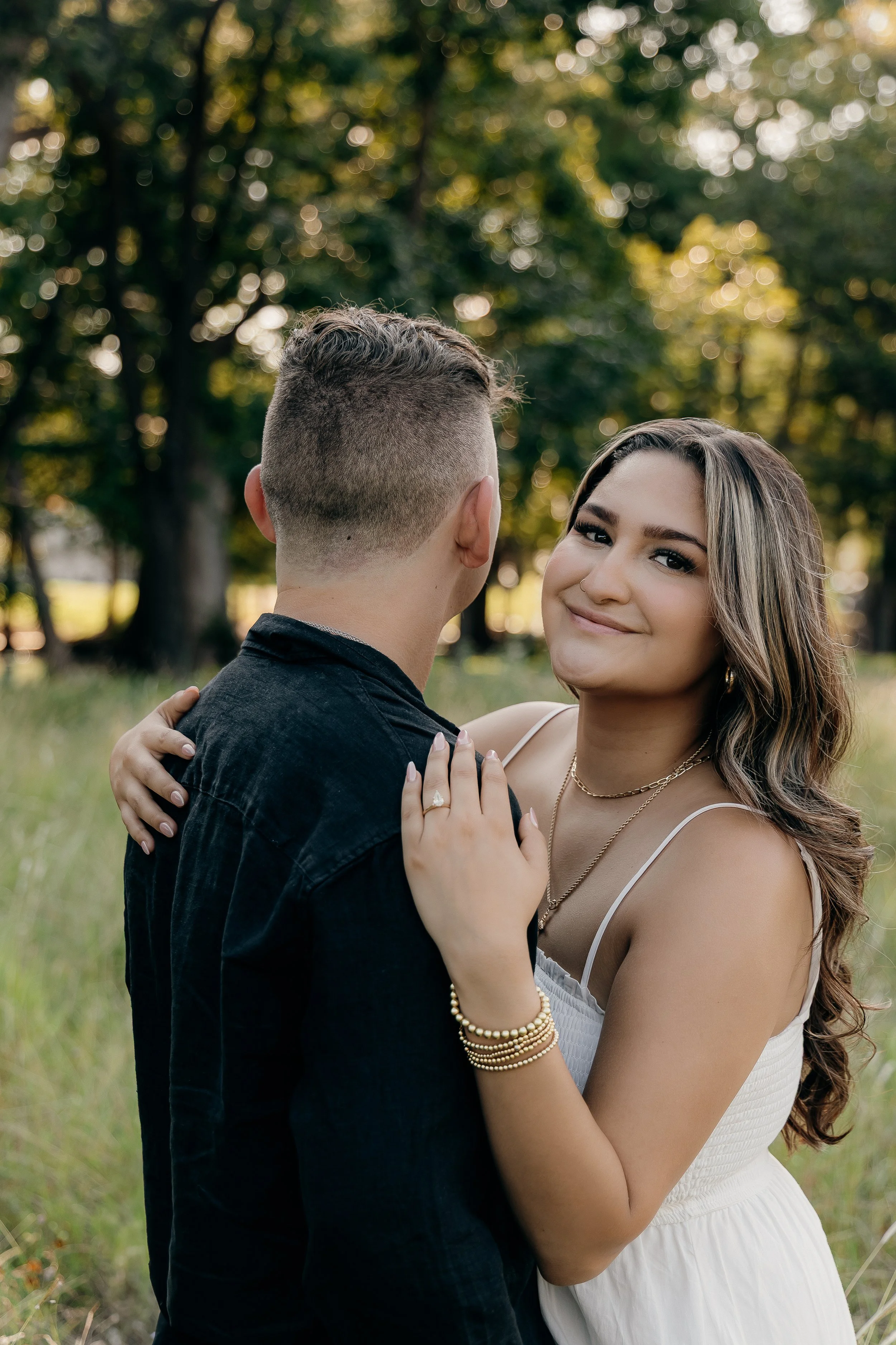 A woman in a white dress and jewelry embraces a man in a black shirt outdoors, with trees and sunlight in the background.