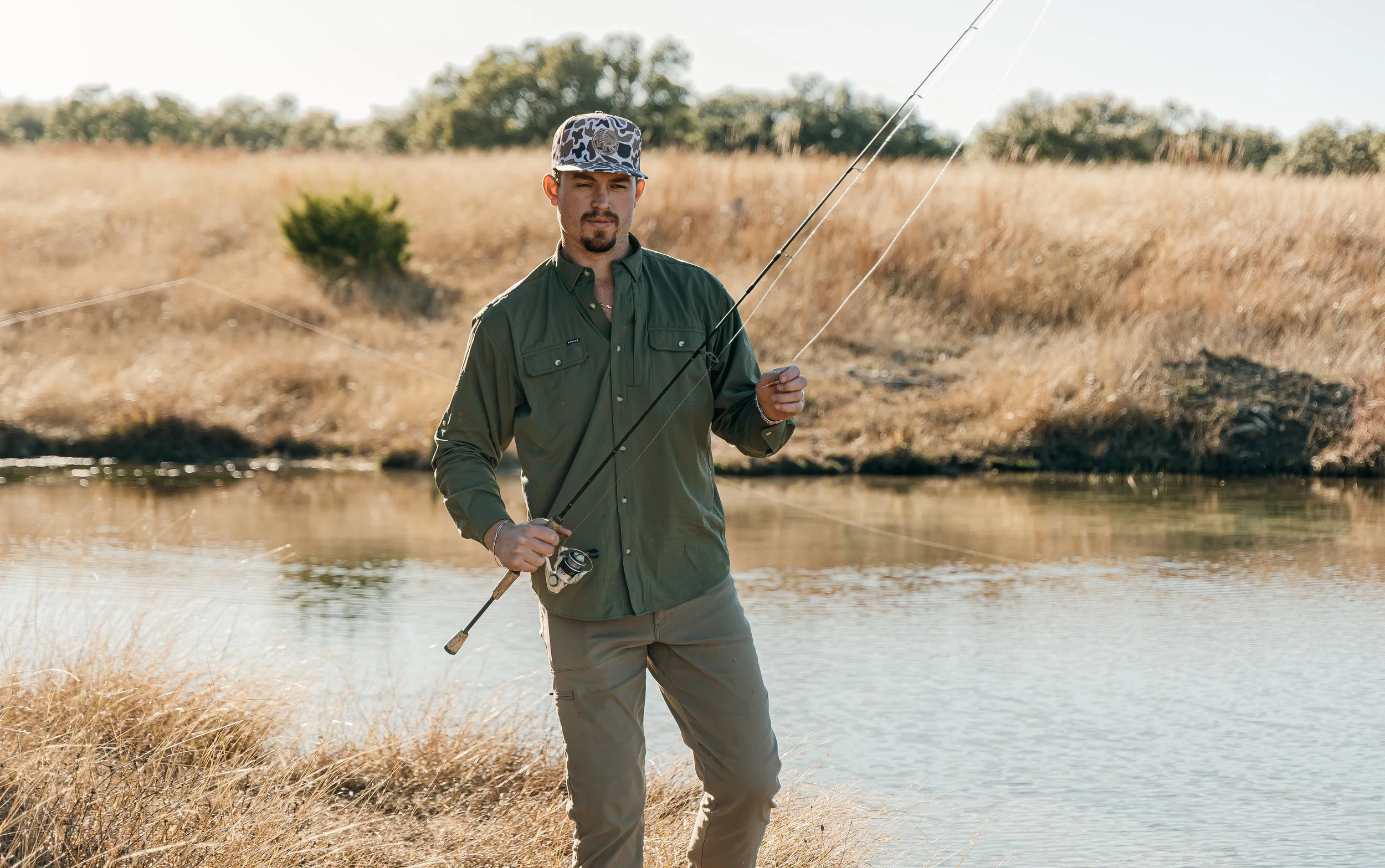 A man standing by a river holding a fishing rod in a natural outdoor setting with tall, dry grass and trees in the background.