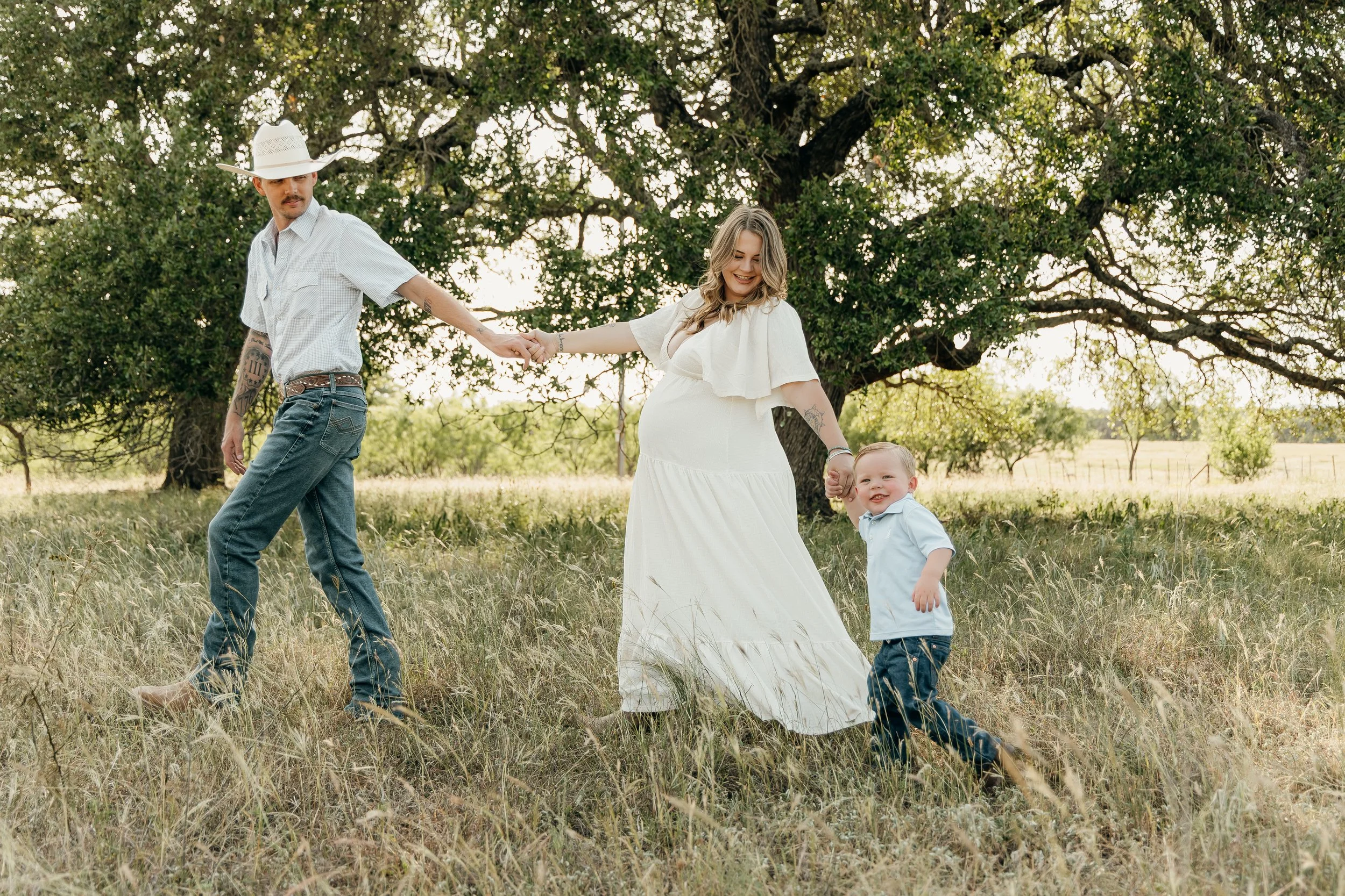 A family of three holding hands and playing in a grassy field with large trees in the background, during a sunny day.