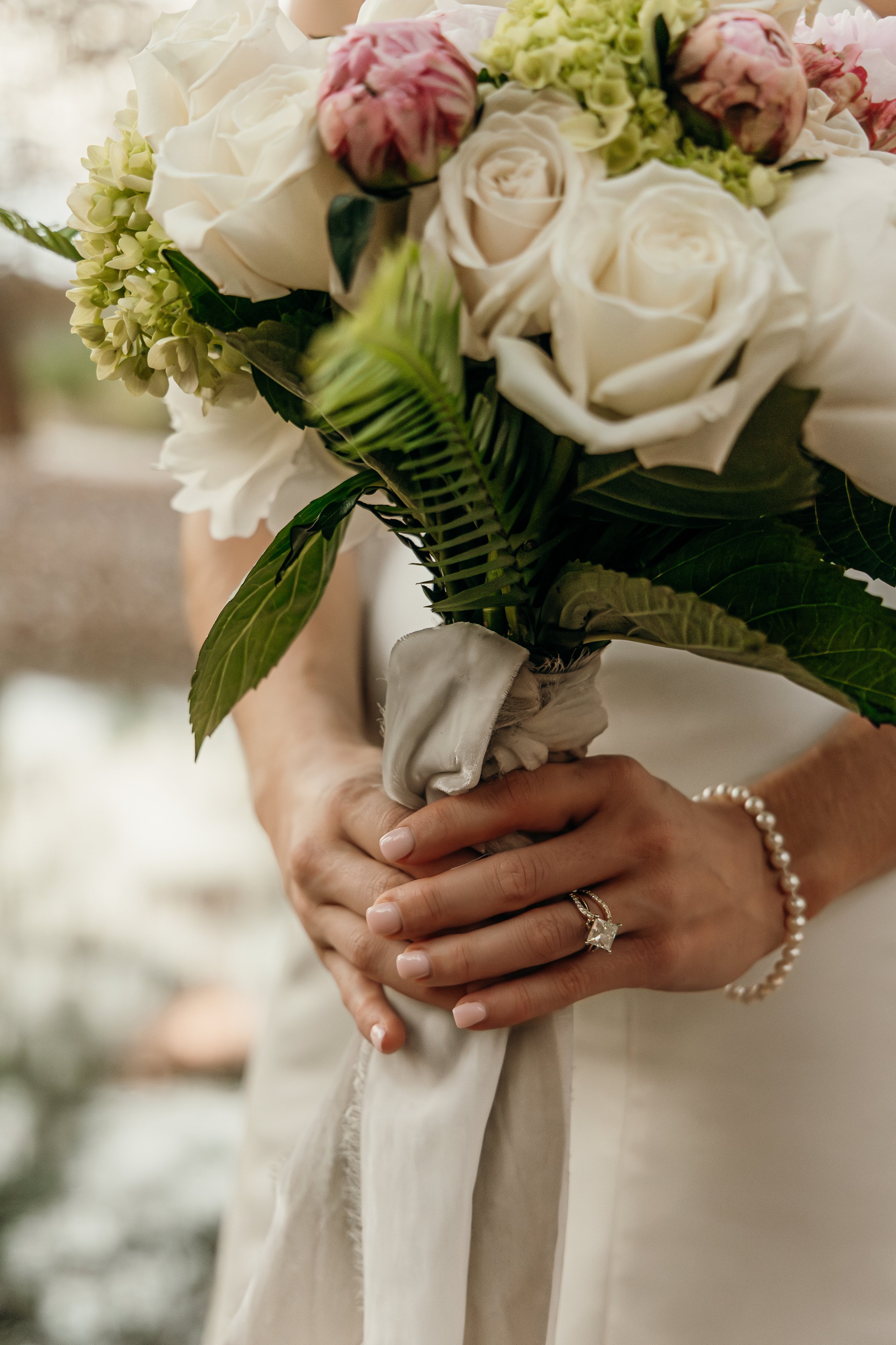 Close-up of a woman holding a bouquet of white roses, pink peonies, and green hydrangeas, with rings and a pearl bracelet visible on her hands.