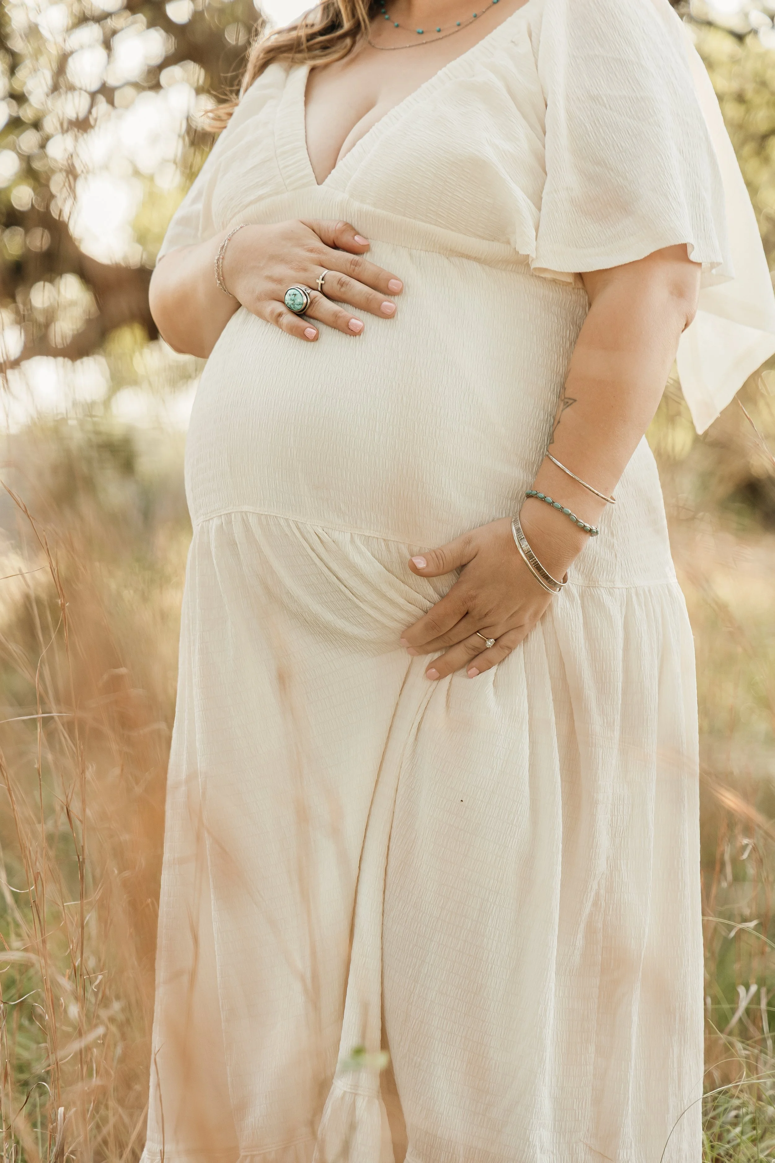 A pregnant woman standing outdoors in a field, wearing a cream-colored loose dress, with her hand resting on her belly and wearing multiple jewelry pieces.