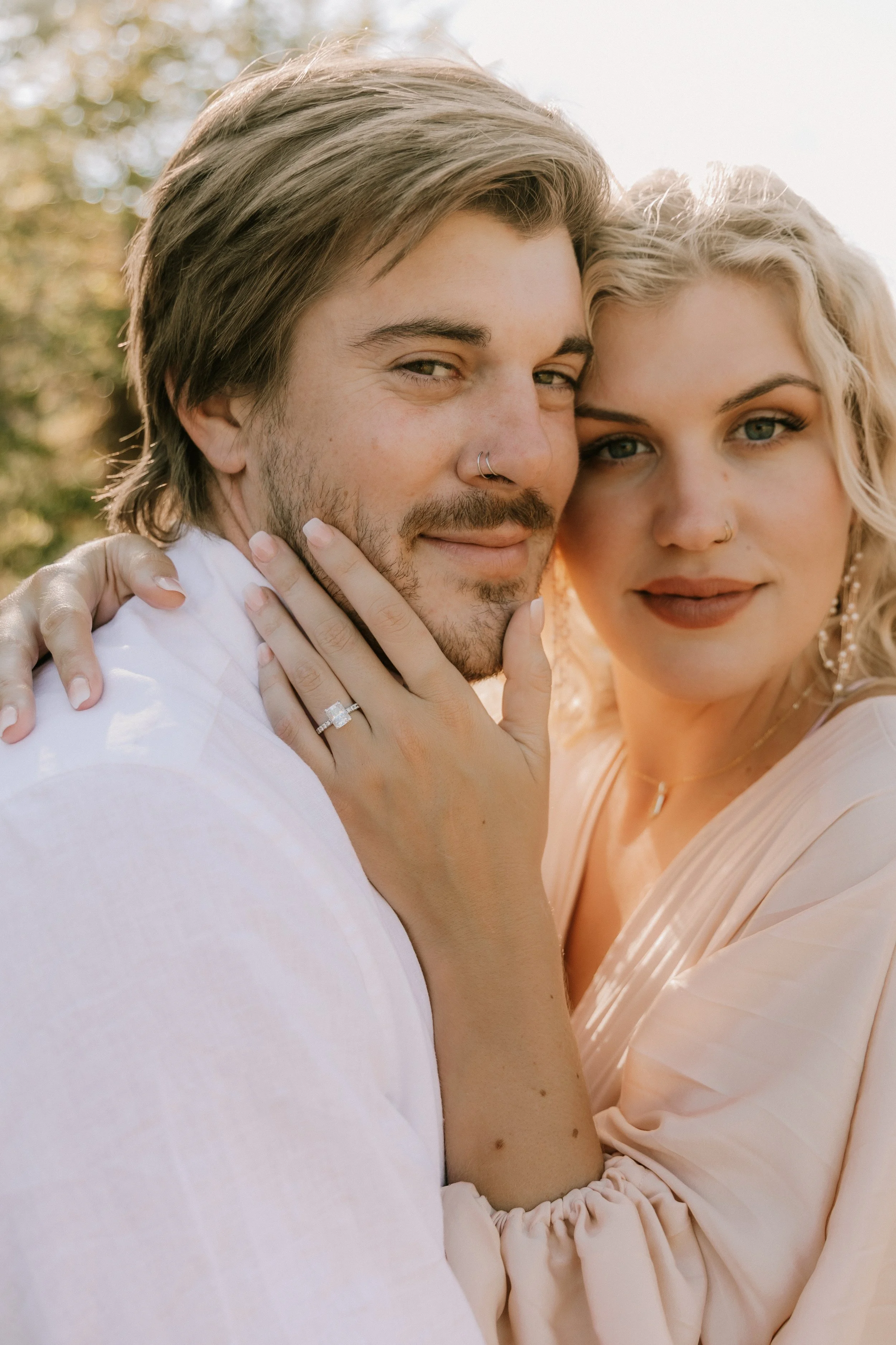 Close-up of a smiling couple embracing outdoors, showing an engagement ring on the woman's finger and their faces close together.