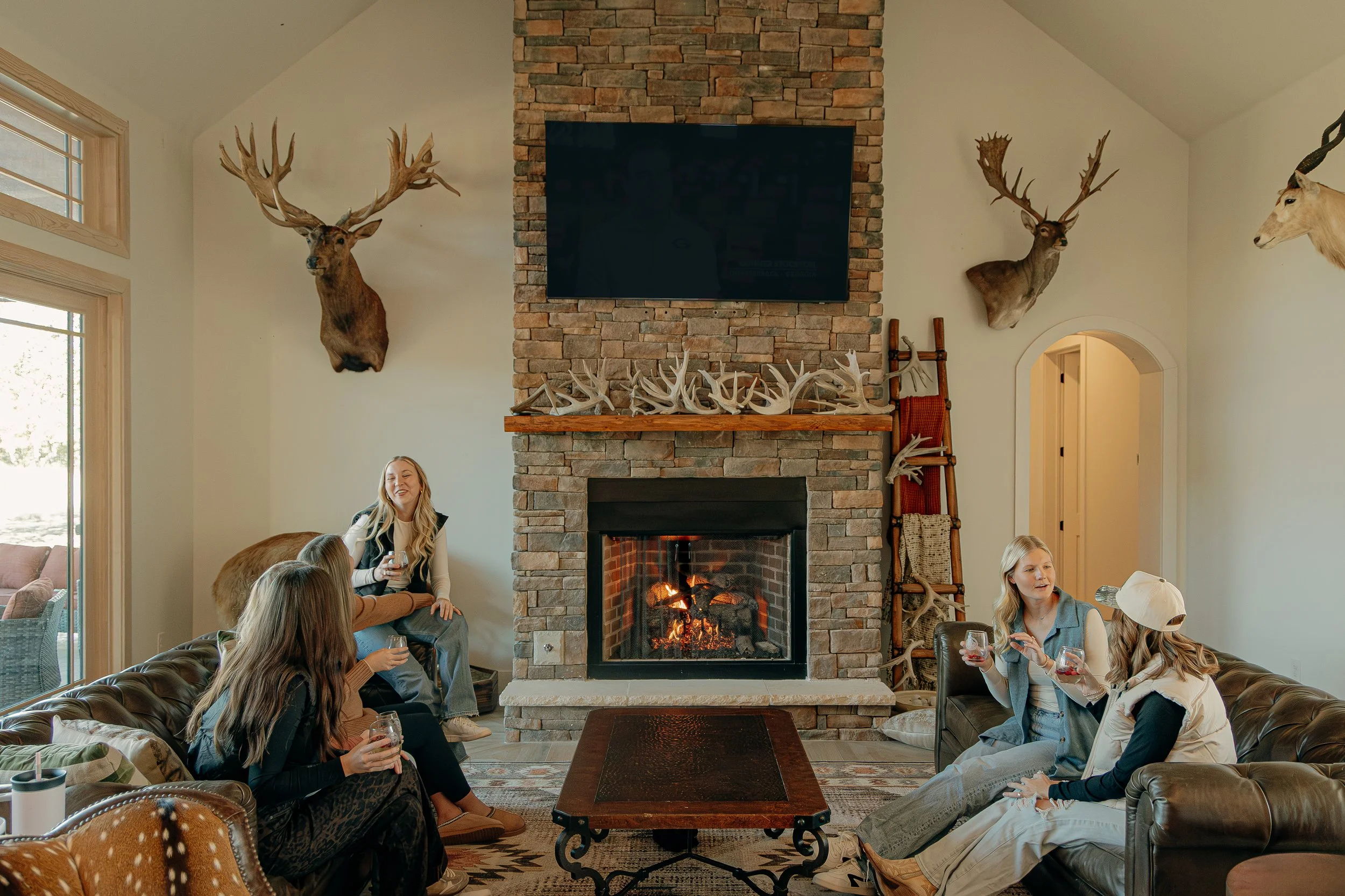 People sitting in a living room with a stone fireplace, deer and elk head mounts on the walls, and a ladder with antlers leaning against the wall.