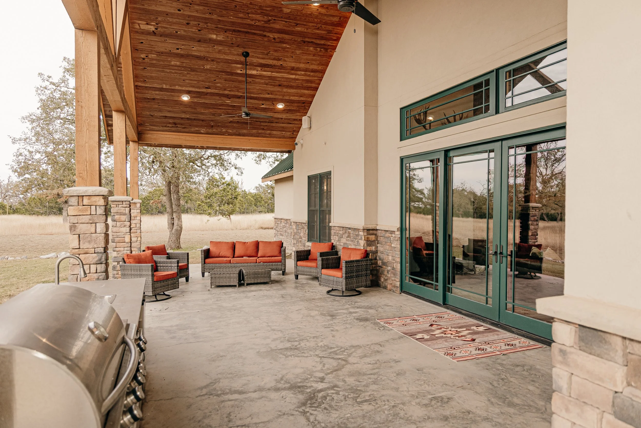 Covered outdoor patio with wicker furniture and orange cushions, stone and stucco house exterior, large glass doors, wood ceiling, and a grill on the left.