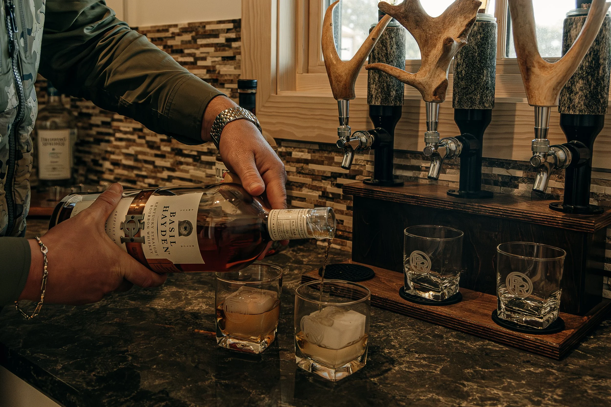 Person pouring Basil Hayden bourbon into a glass with ice on a dark marble countertop, with two other glasses and a wooden liquor dispenser in the background.