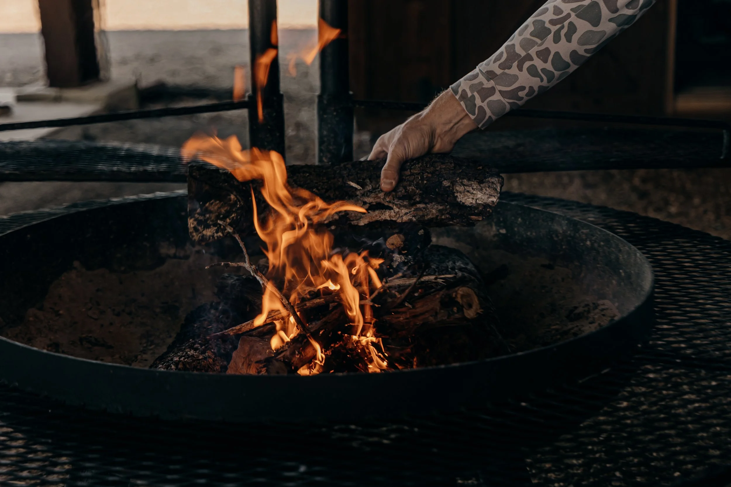 Person placing a log into an open fire pit with flames burning around the logs.