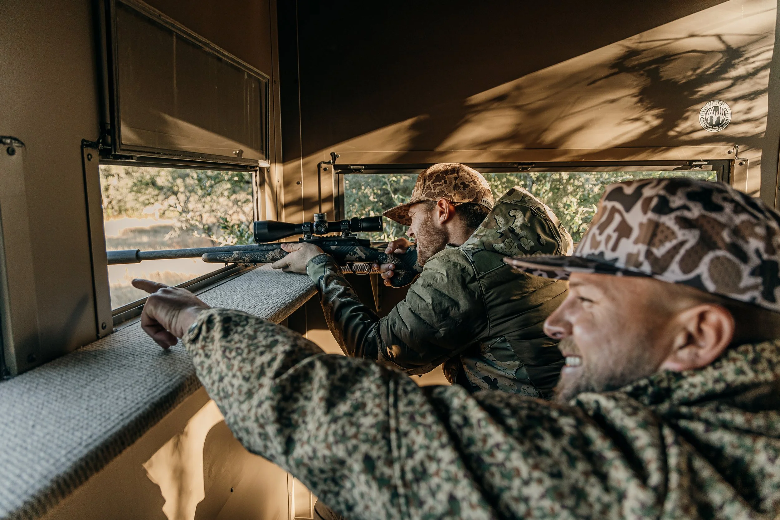 Two soldiers inside a mobile sniper hide, one aiming through a scoped rifle and the other observing, both in camouflage gear.