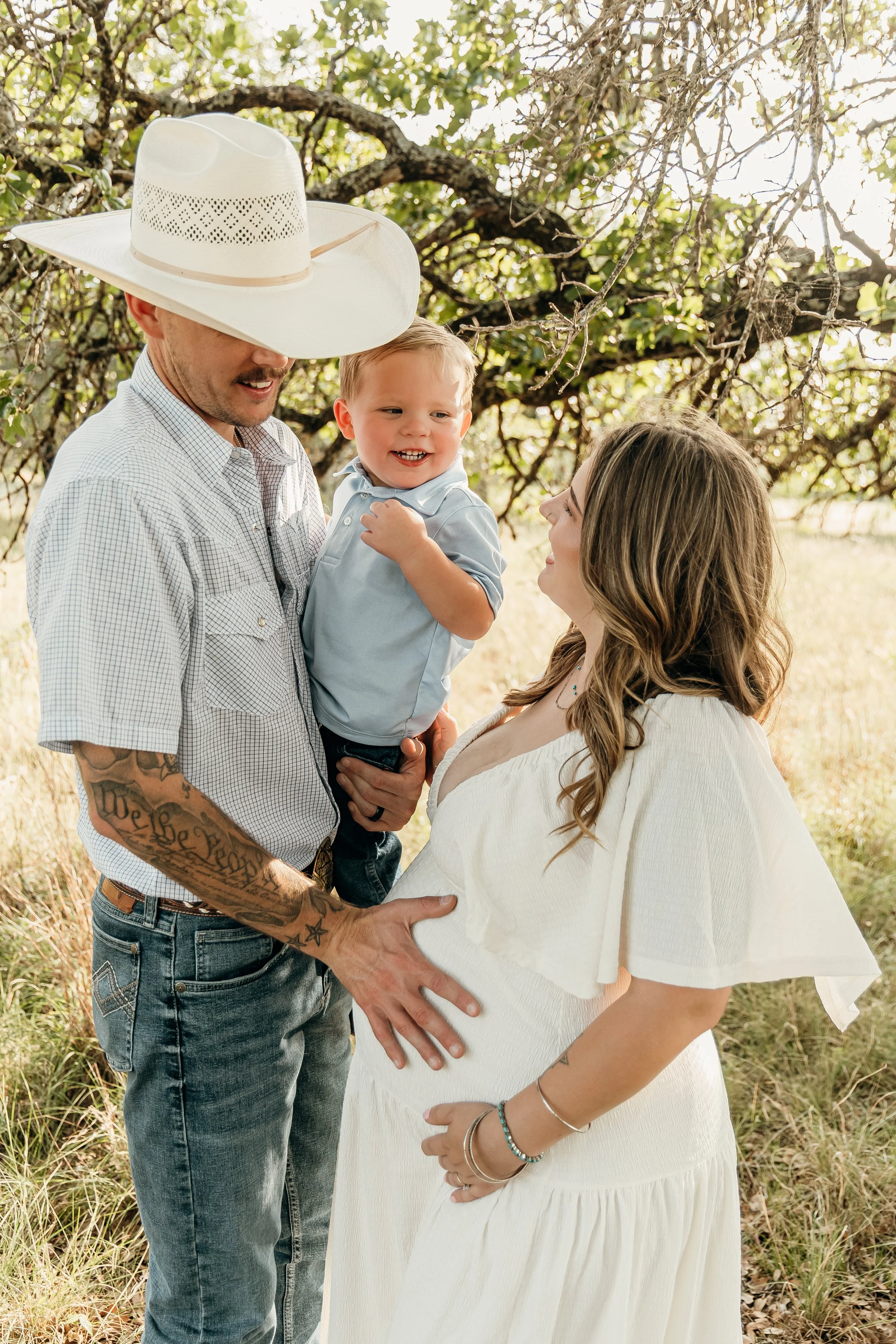 A family outdoors, a man in a cowboy hat holding a smiling young boy, woman looking at the boy and touching her pregnant belly, under a tree with sunlight filtering through.