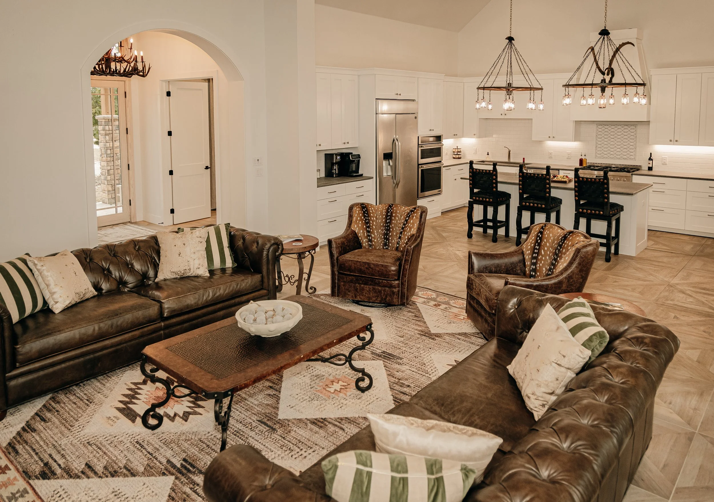 Living room with leather sofas and armchairs, a patterned area rug, and a wooden coffee table. The open kitchen has white cabinets, an island with barstools, and modern appliances. Pendant lights hang above the kitchen area.