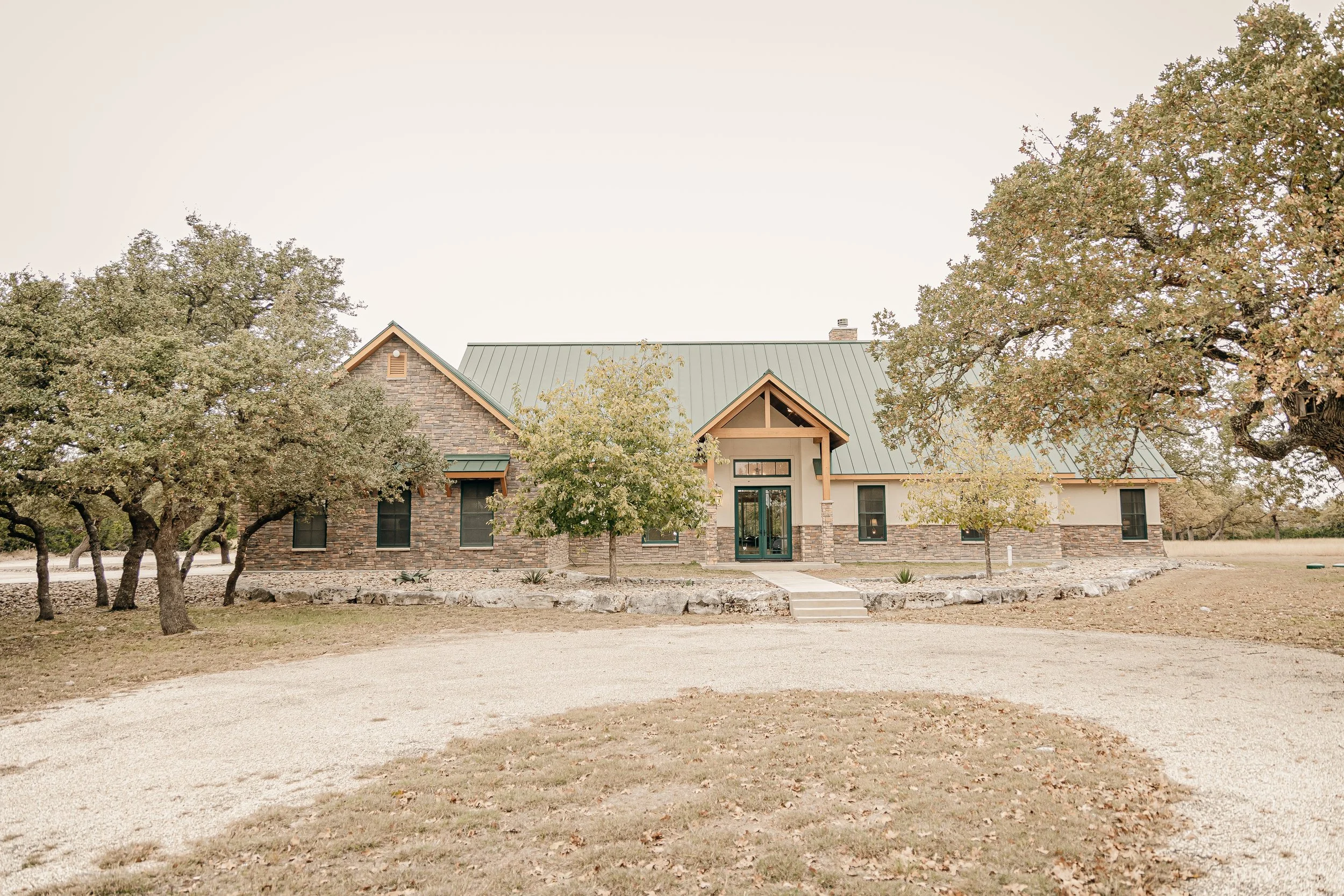 Front view of a large modern house with a green metal roof, stone and stucco exterior, set amidst trees on a gravel driveway