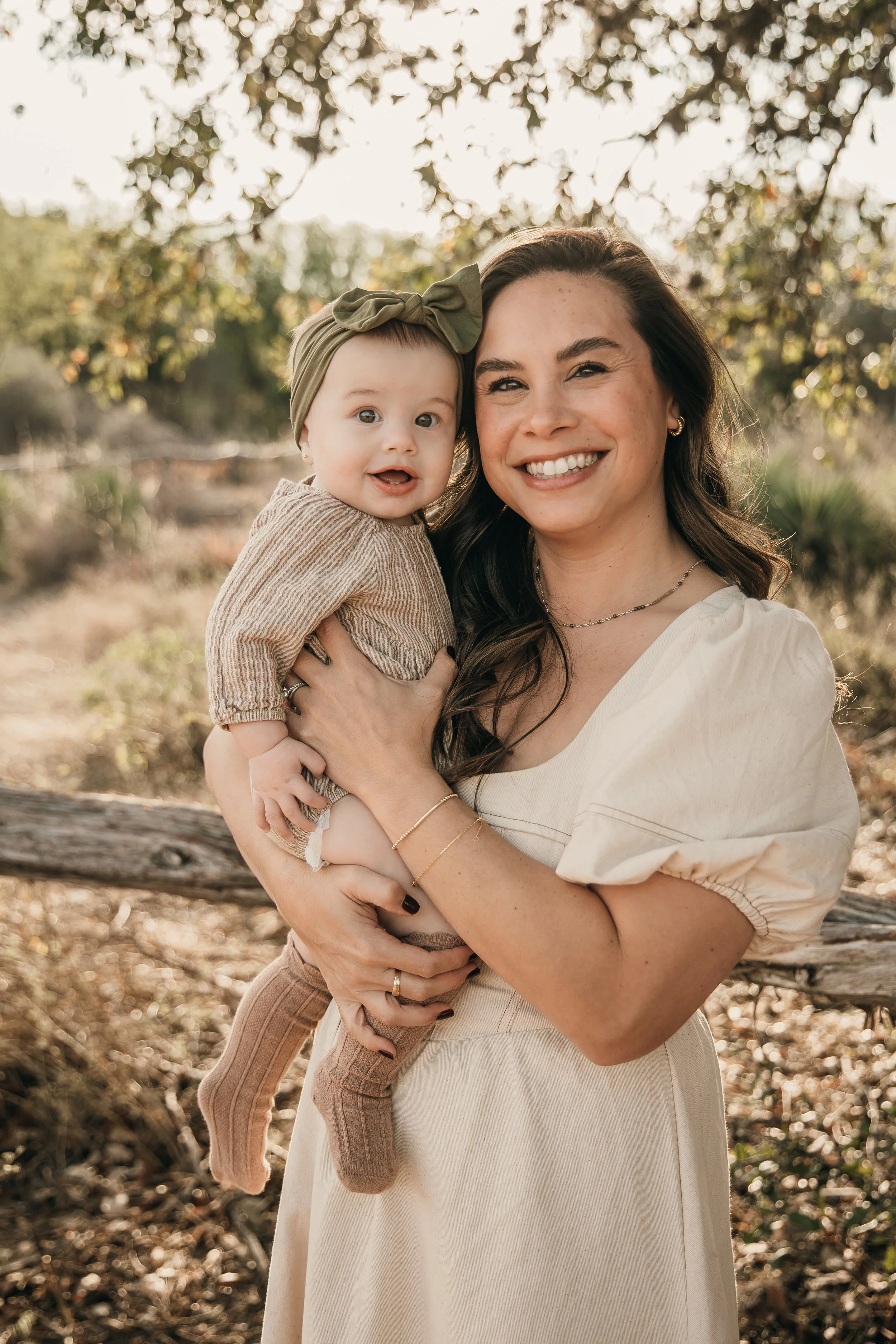 A woman holding a baby girl outdoors with trees and sunlight in the background.