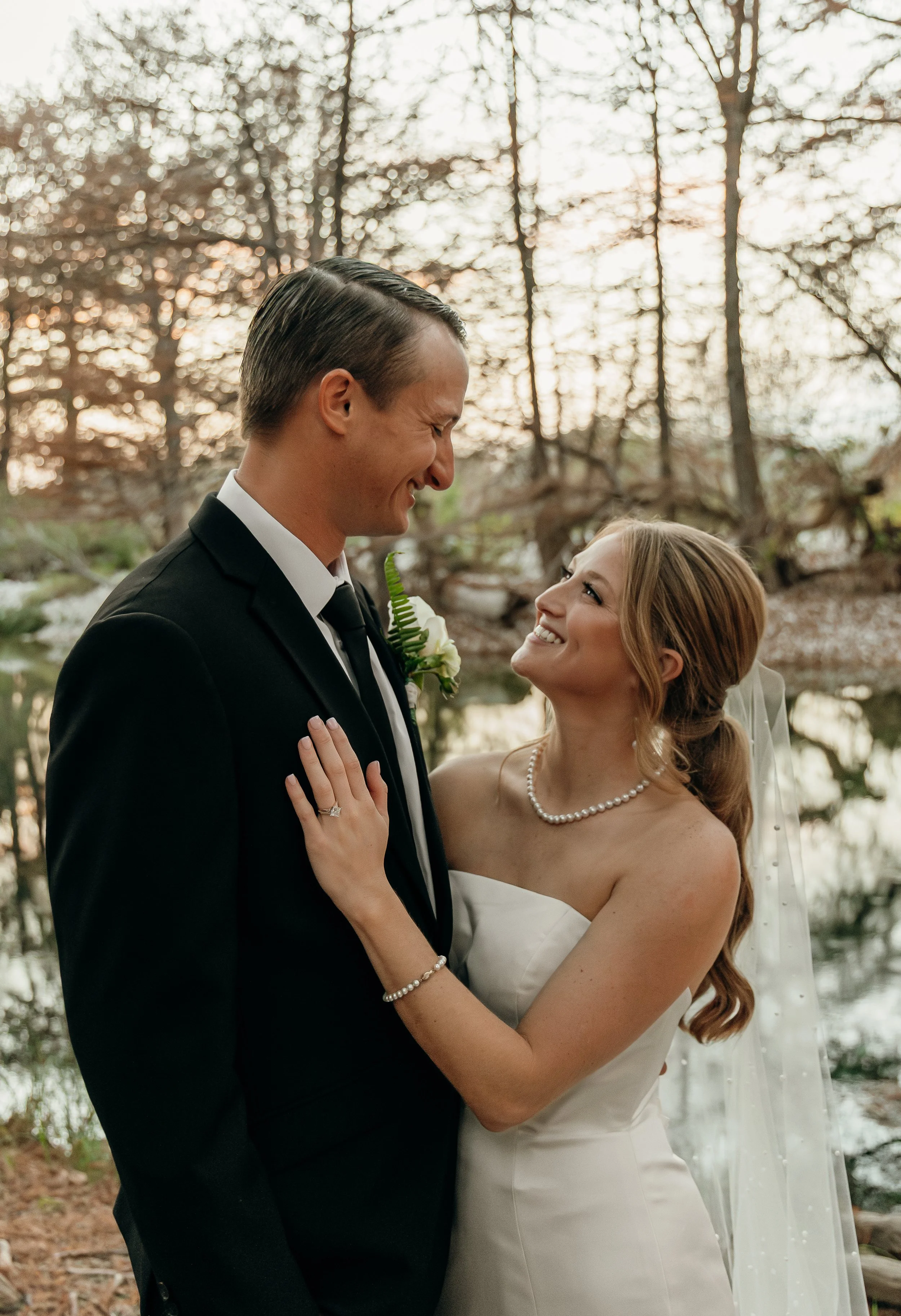 A bride and groom smiling at each other outdoors during sunset, surrounded by trees and a pond.
