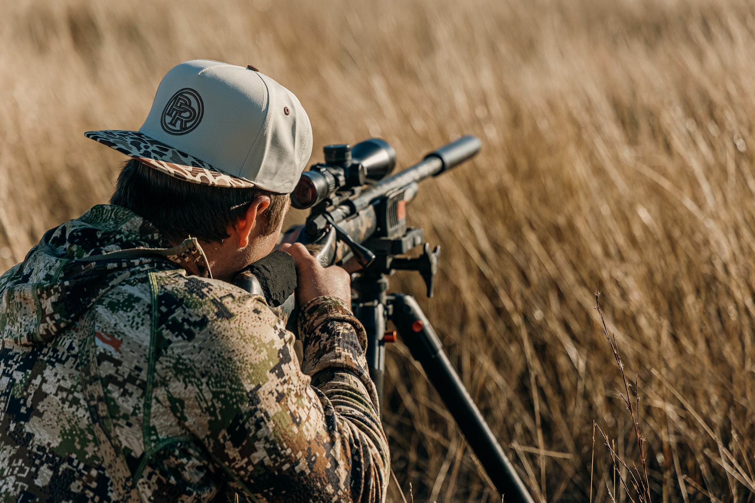 Person in camouflage clothing aiming a scoped rifle in a field of tall grass or wheat.