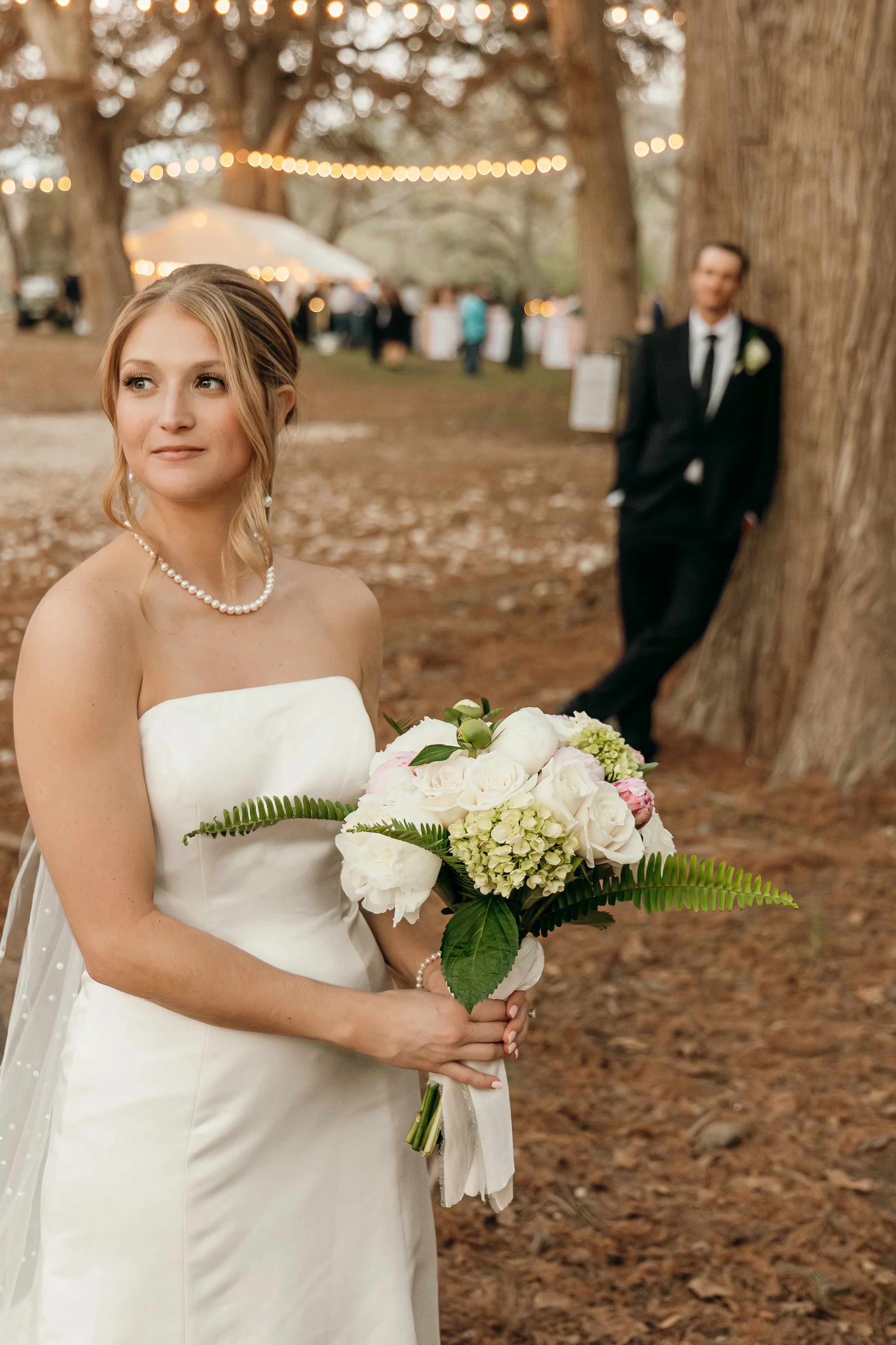 Bride holding bouquet of white and pink flowers with greenery, standing outdoors in a wooded area, groom leaning against a large tree in the background, with string lights and tables in the distance.