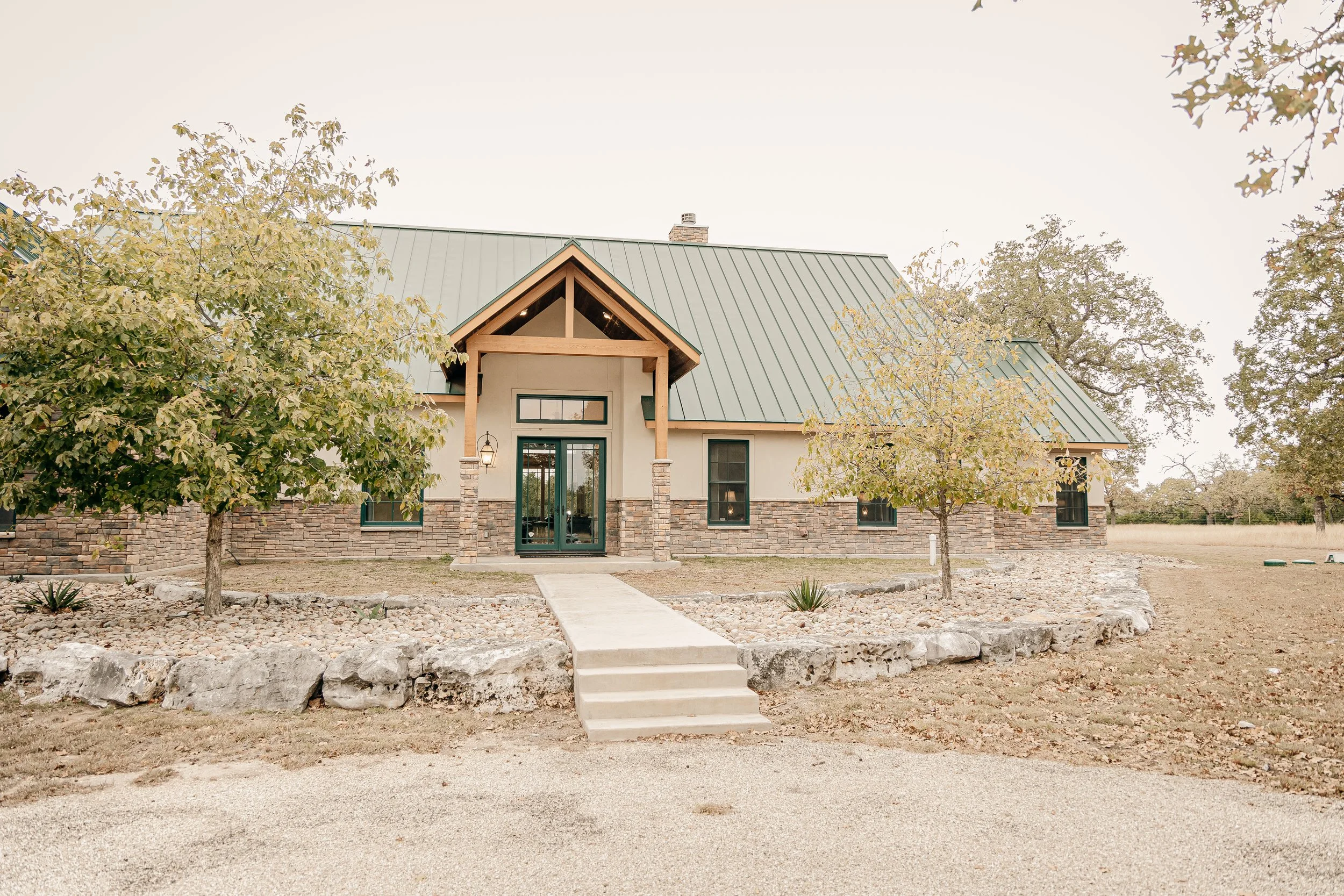 Front view of a house with a metal green roof, stone accents, and a concrete pathway leading to the front door, surrounded by trees and a gravel yard.