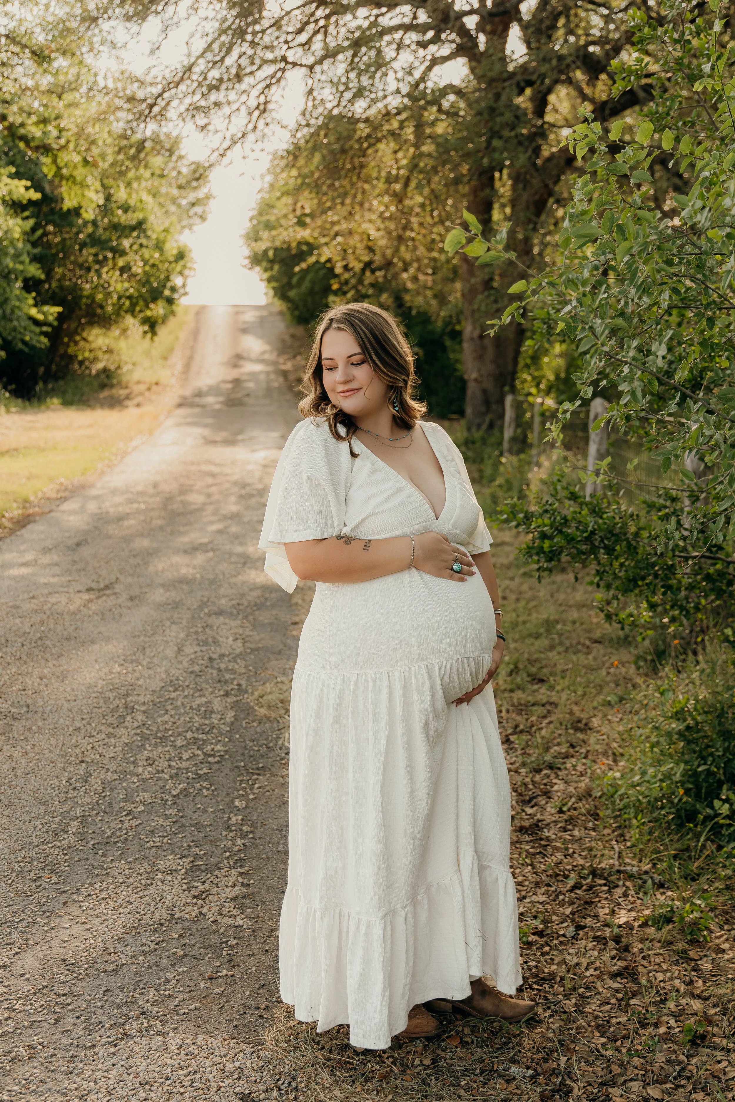 A pregnant woman standing on a rural dirt road surrounded by trees, smiling and gently holding her baby bump in the late afternoon sunlight.
