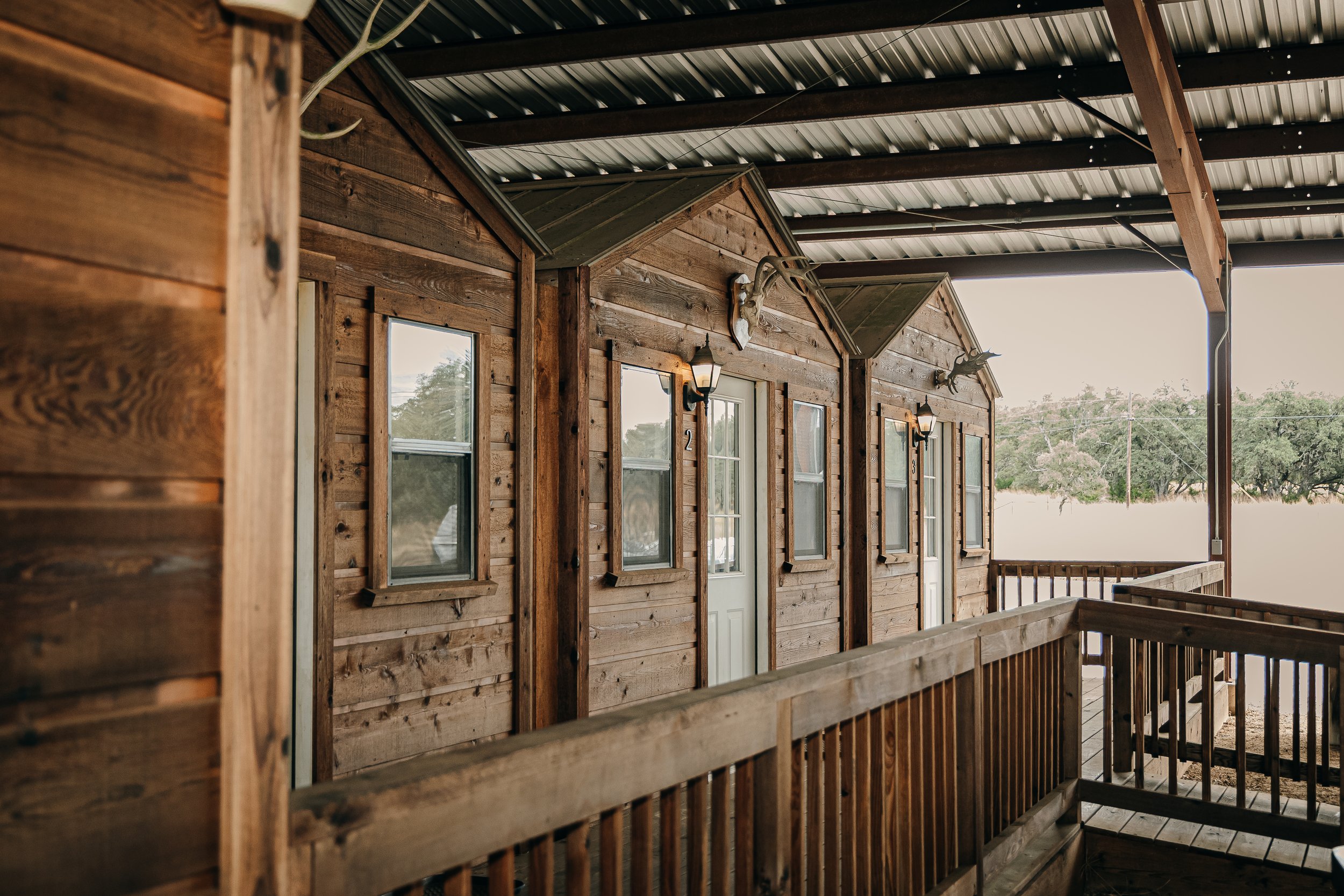Wooden cabins on a porch with a metal roof, decorated with animal skull and antlers, set in a rural area with trees in the background.