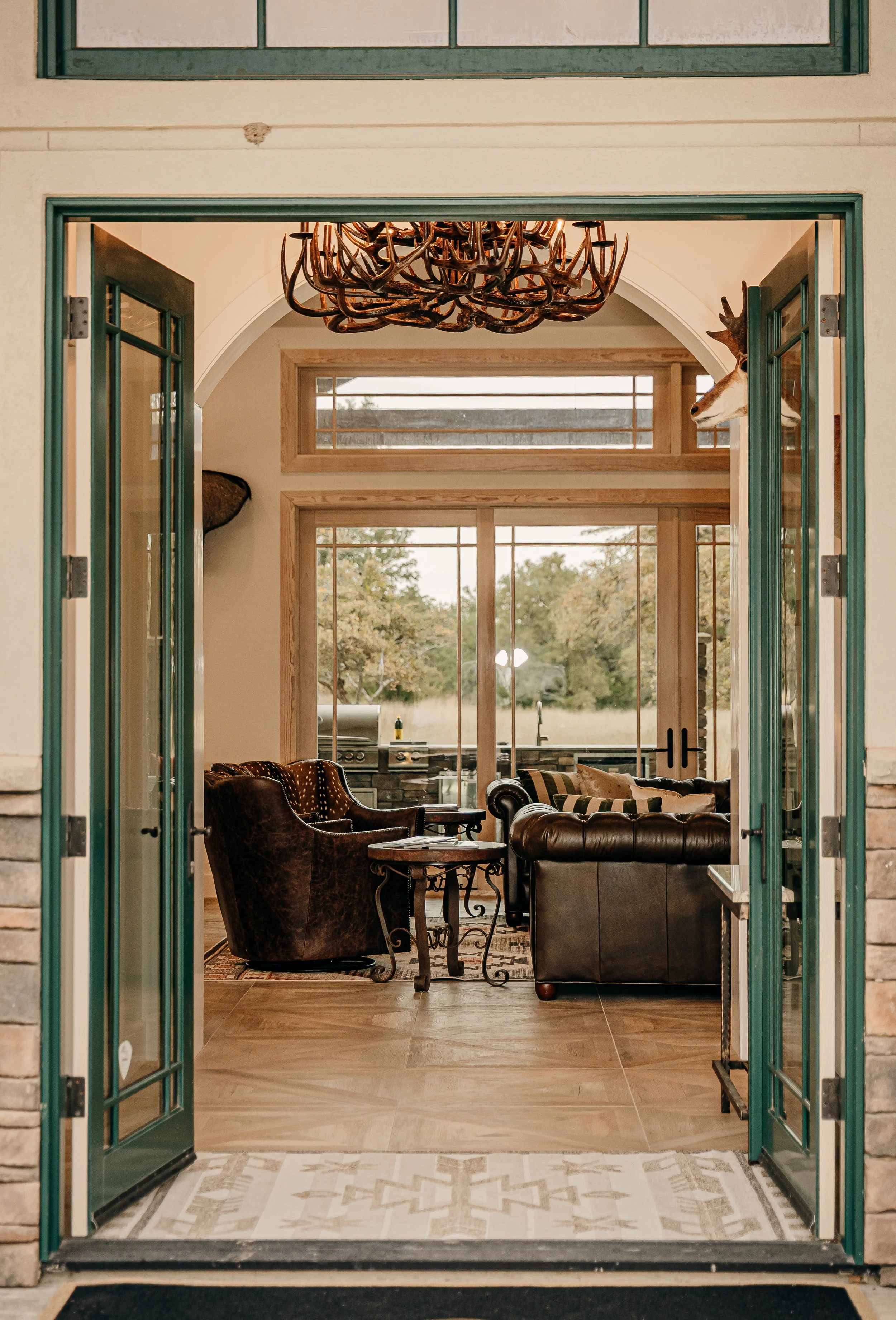 Interior view through open porch doors of a living room with leather couches, a rustic chandelier, large windows, and a mounted deer head on the wall.
