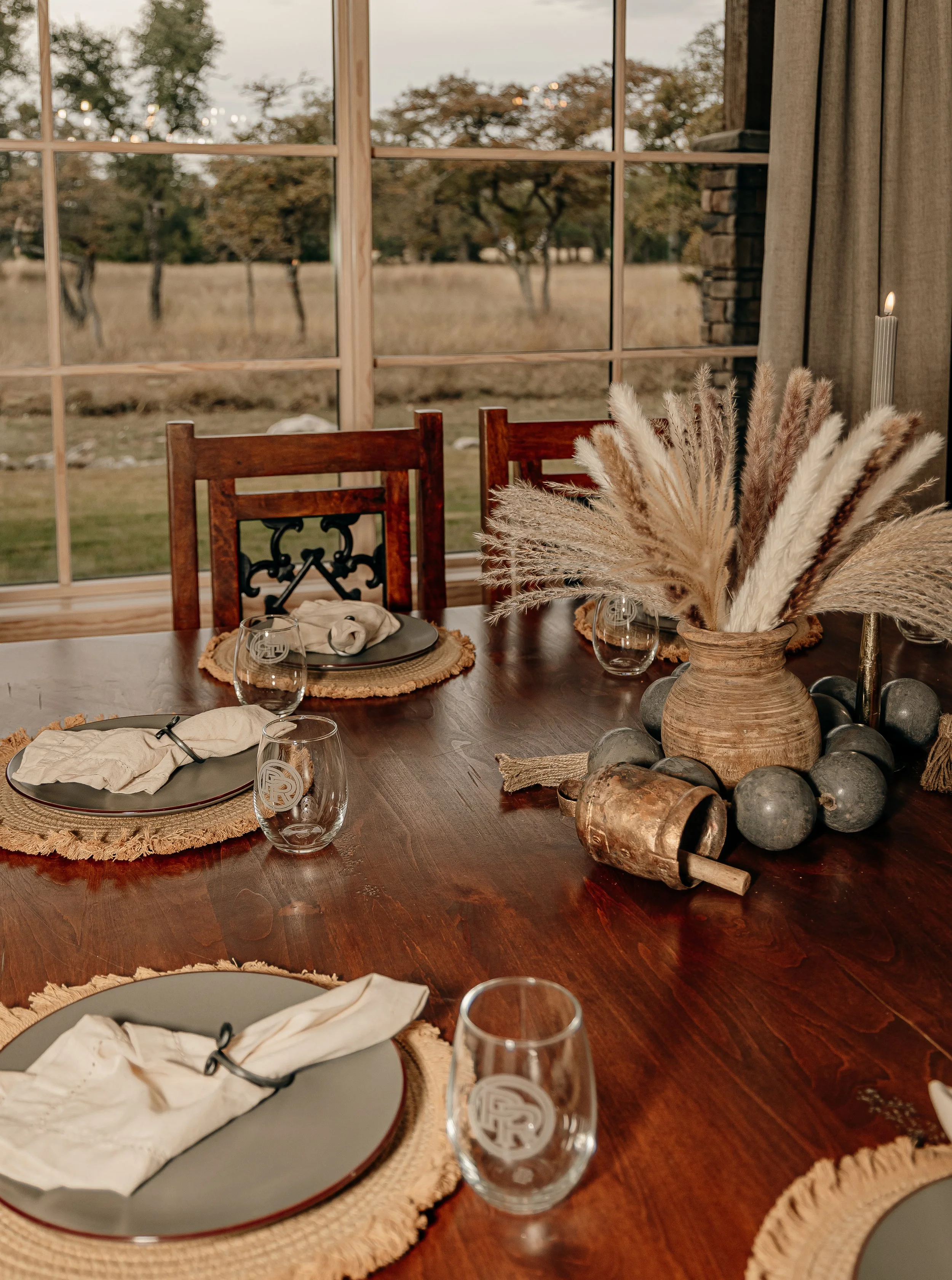 A rustic dining table set for four, with woven placemats, gray plates, cloth napkins wrapped with black rings, and clear glasses featuring a logo. The centerpiece is a large clay vase filled with pampas grass, surrounded by decorative stones and smal