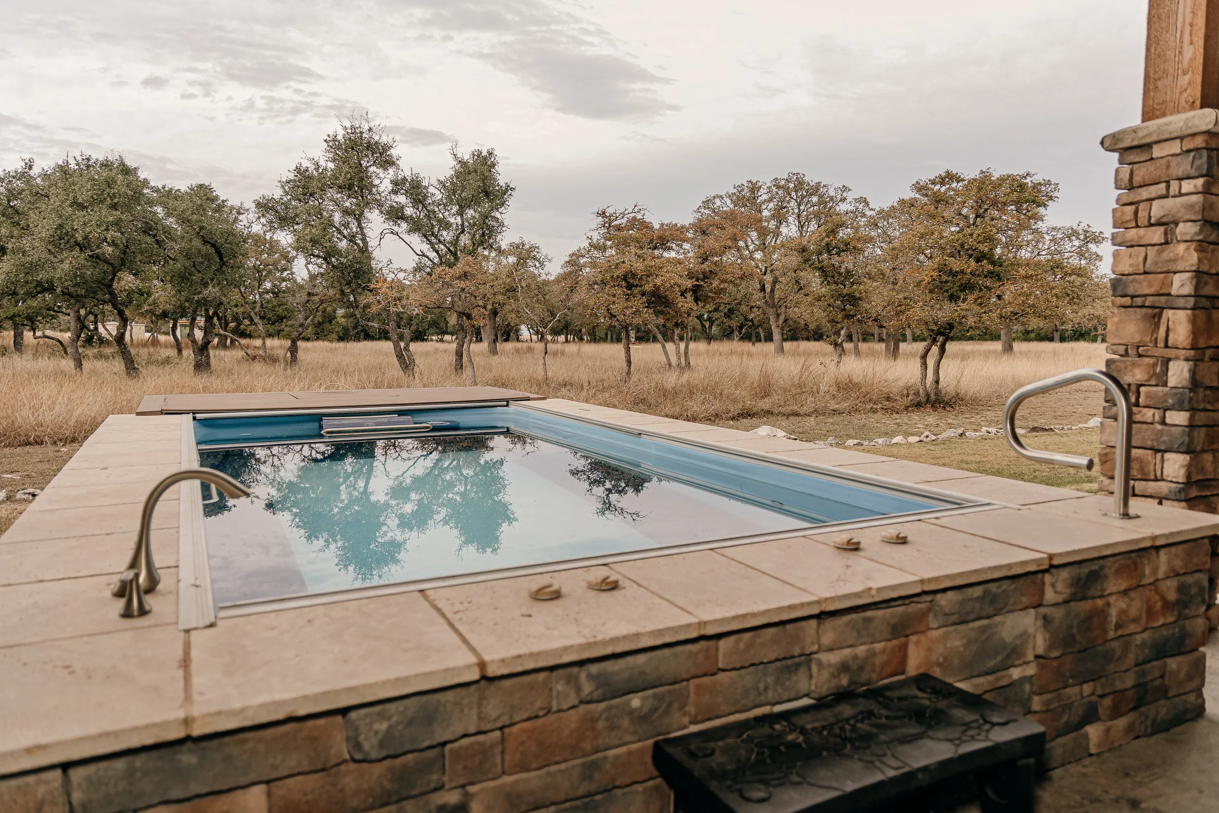 A backyard hot tub surrounds with stone tiles, set against an open field with trees and grass, under an overcast sky.