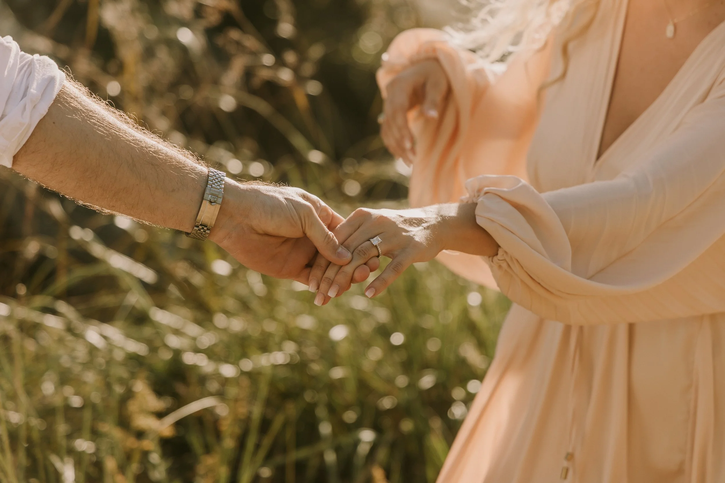 A man and woman holding hands outdoors, with the woman wearing an engagement ring, surrounded by tall grass or plants.
