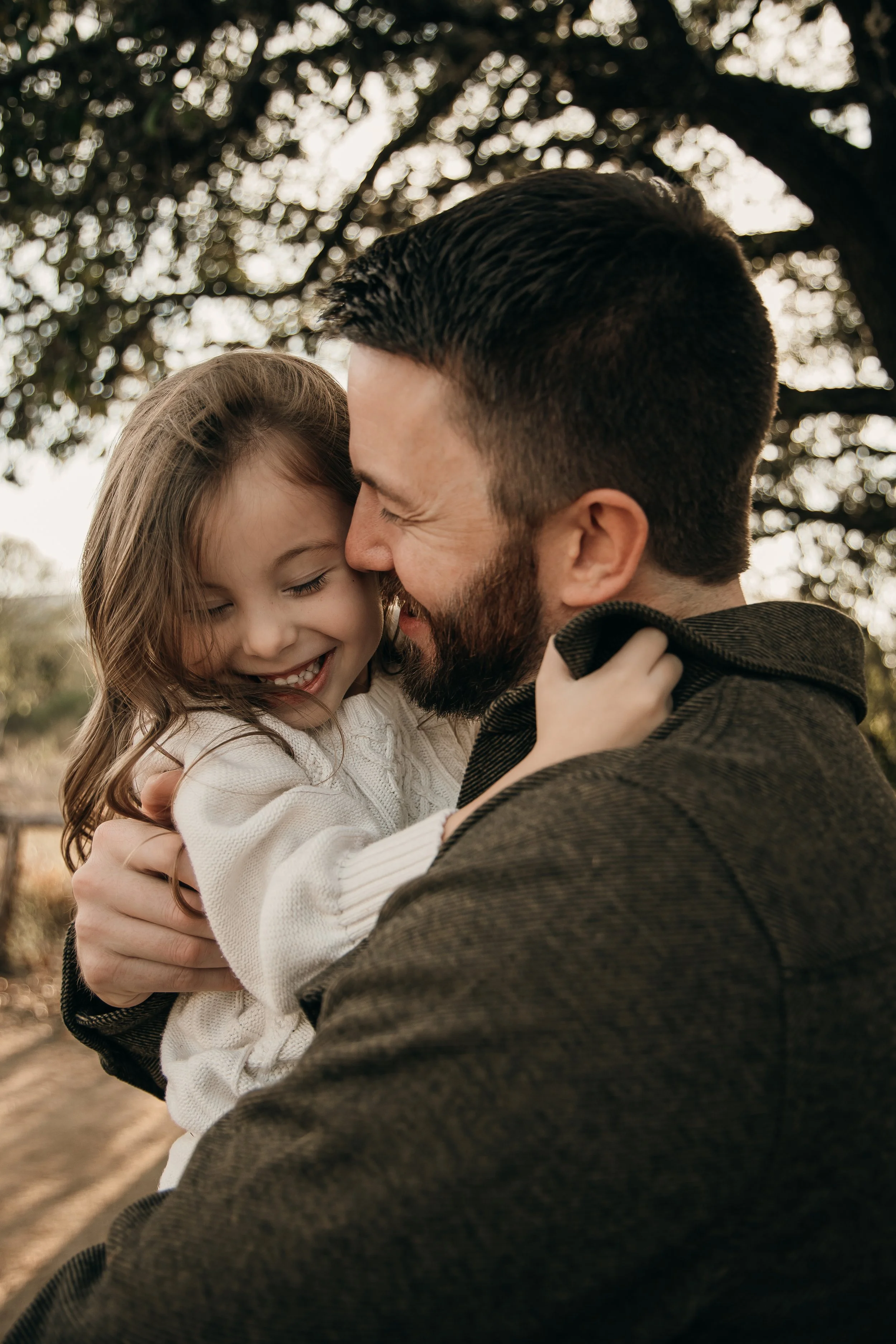 A man and a young girl are smiling and holding each other outdoors, with trees in the background.