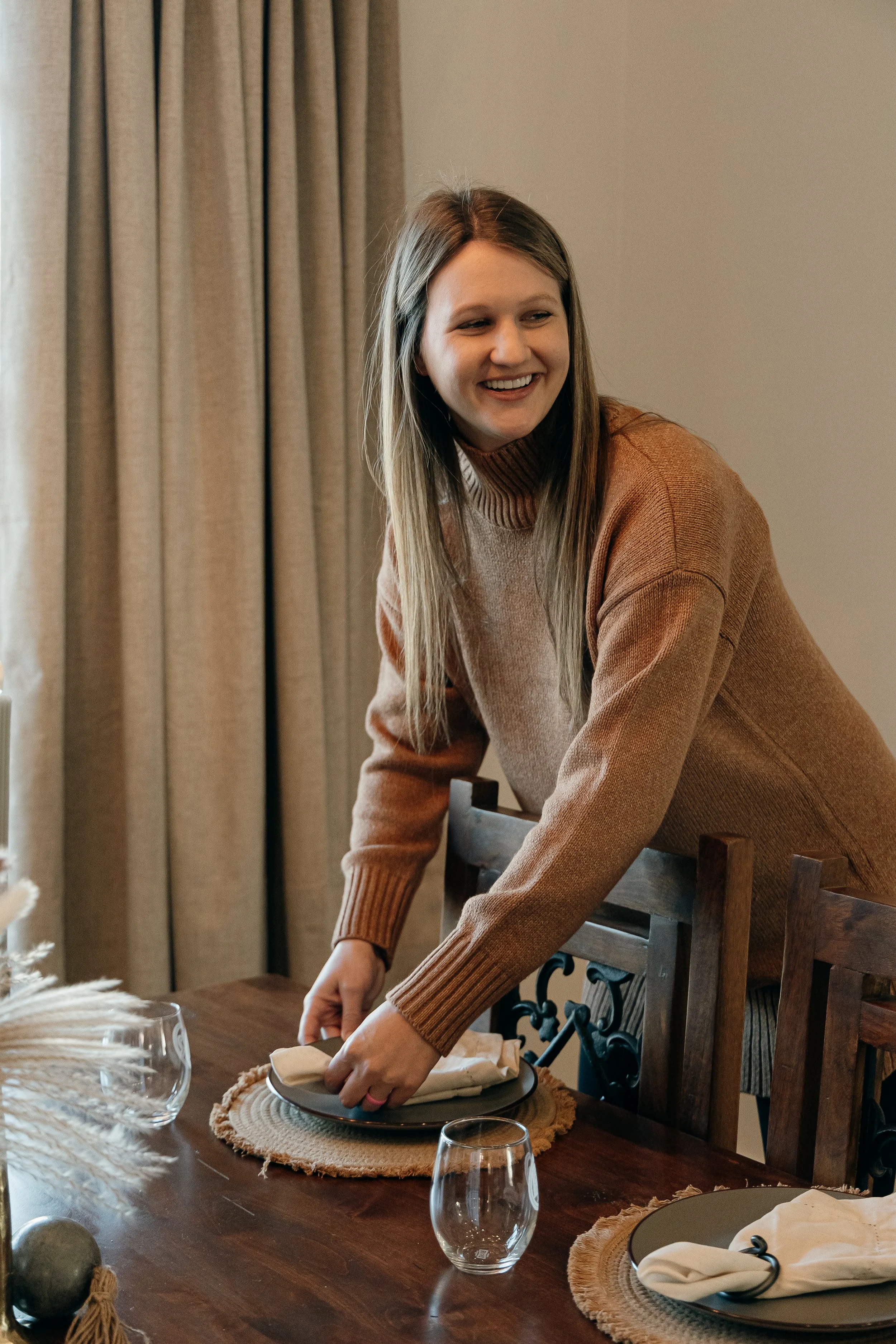 A woman with long hair, wearing a brown sweater, is setting a dining table with plates and glasses in a cozy, well-lit room.