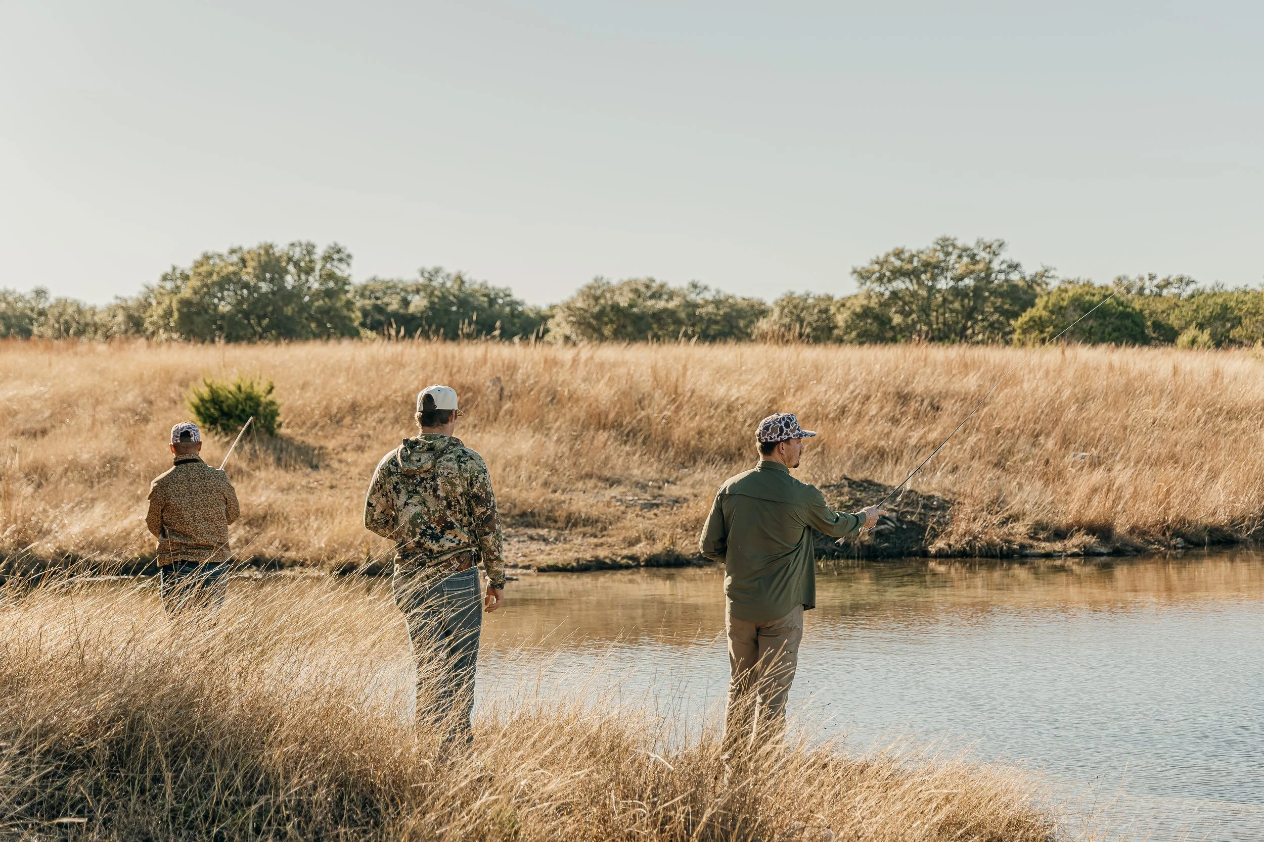 Three men fishing by a river in a grassy field with trees in the background during daylight.
