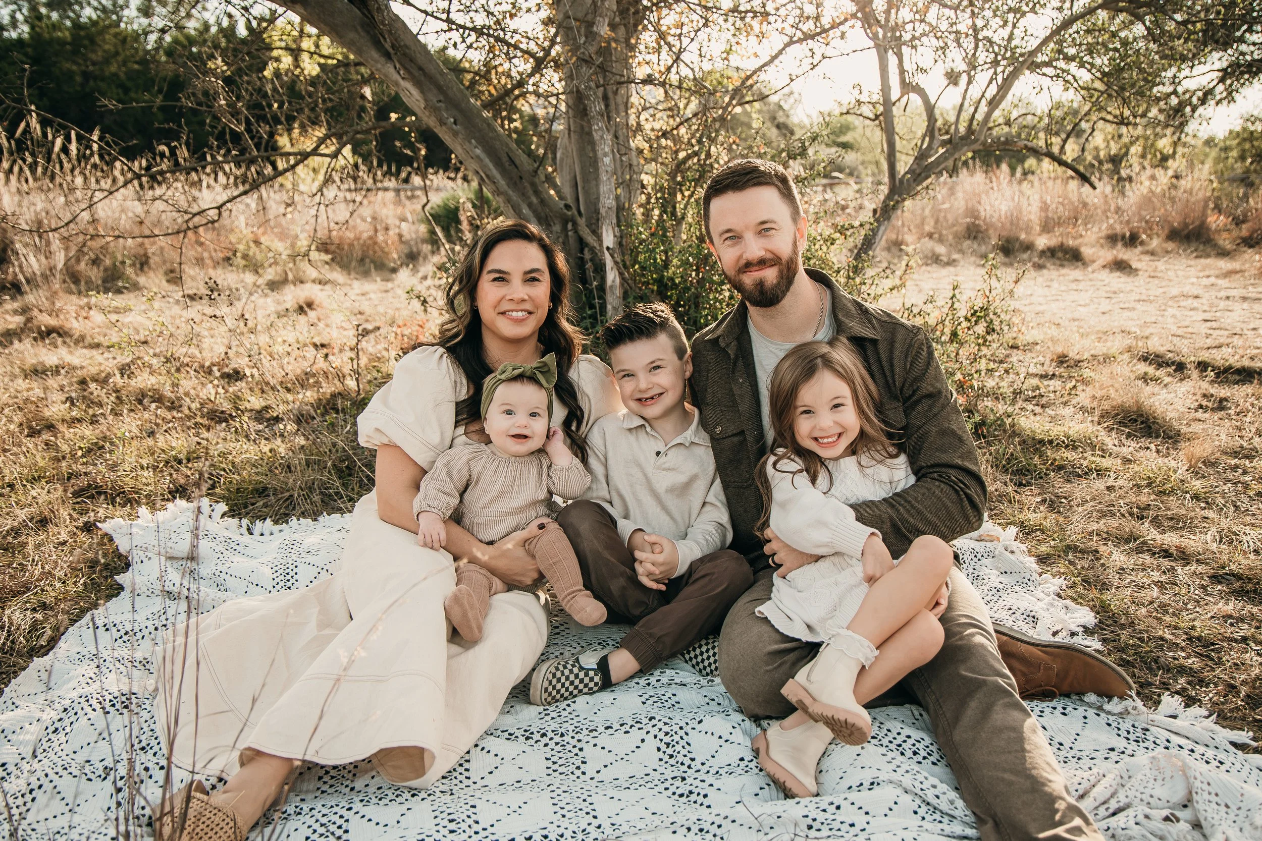 A family of five sitting on a blanket outdoors in a natural setting with trees and dry grass, smiling at the camera.
