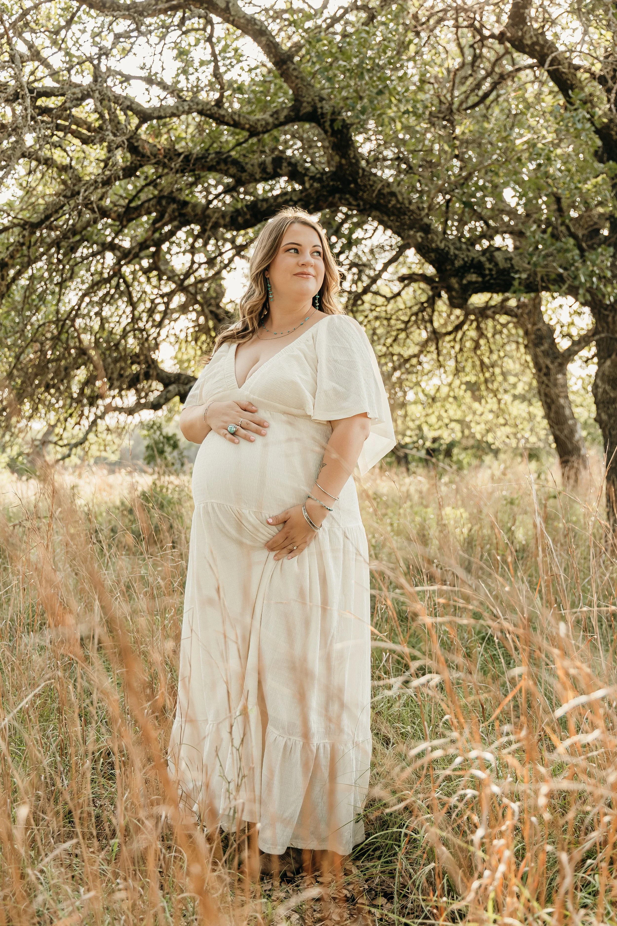 A pregnant woman standing in a field of tall grass beside a large tree, wearing a light-colored, flowing dress, gazing off into the distance.
