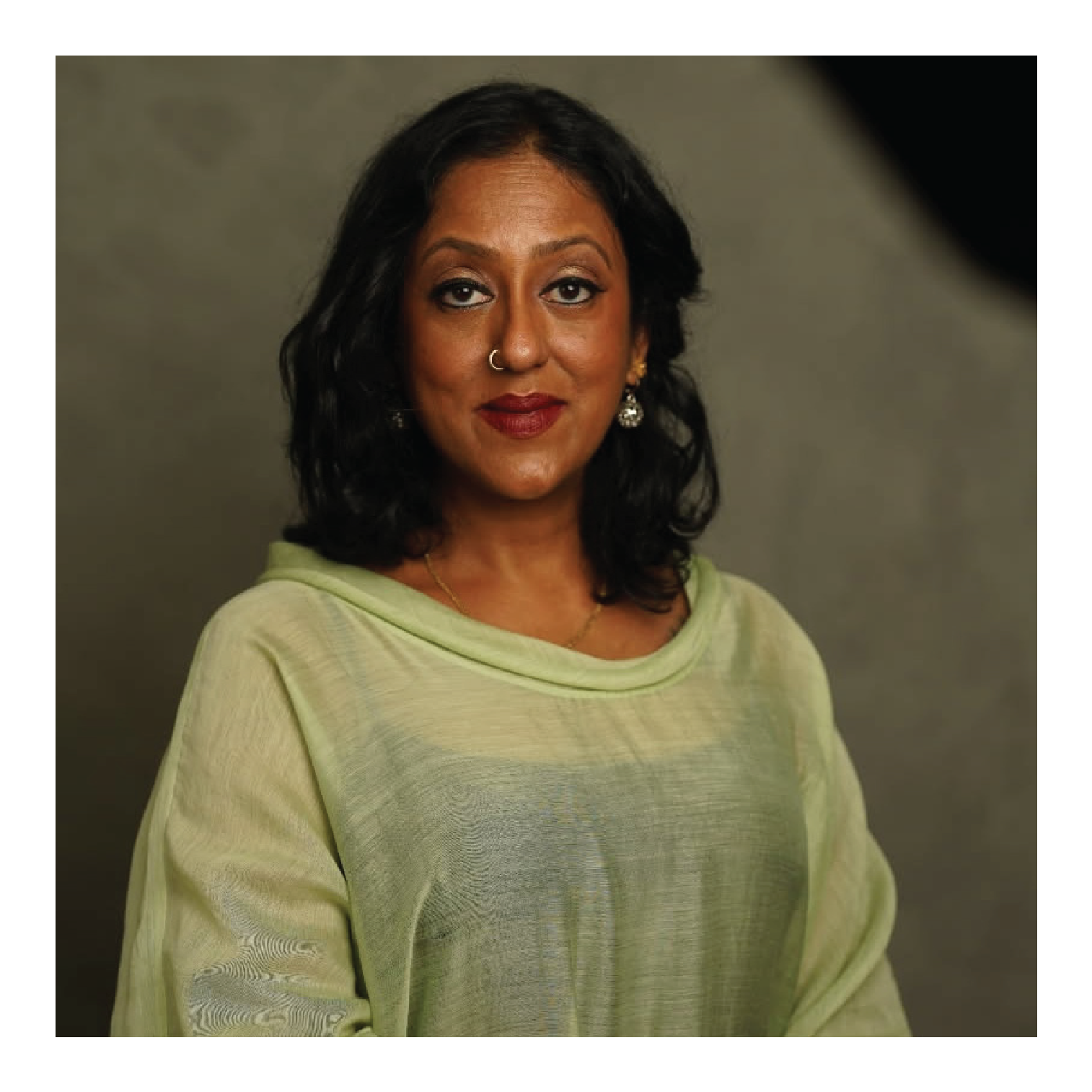 A woman with medium skin tone, black curly hair, gold earrings, and a nose piercing, wearing a light green sweater, standing against a gray background.