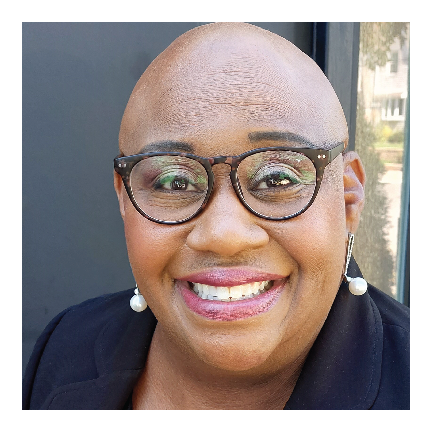 A smiling African American woman with a shaved head, wearing glasses, pearl earrings, and a black blazer, standing outdoors with a blurred background.