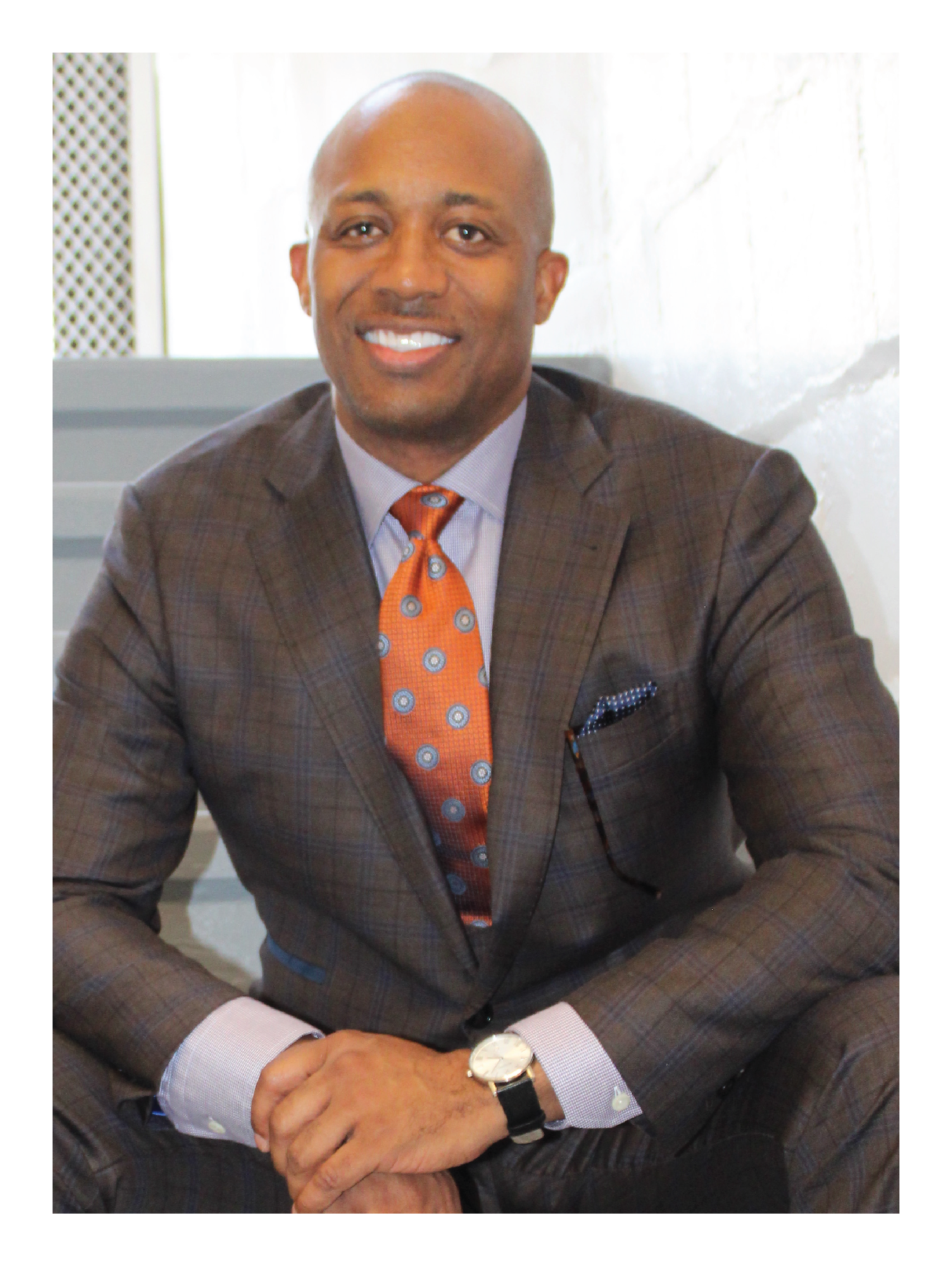 A man in a brown plaid suit with a light shirt and orange patterned tie, sitting indoors with a bright background, smiling at the camera.