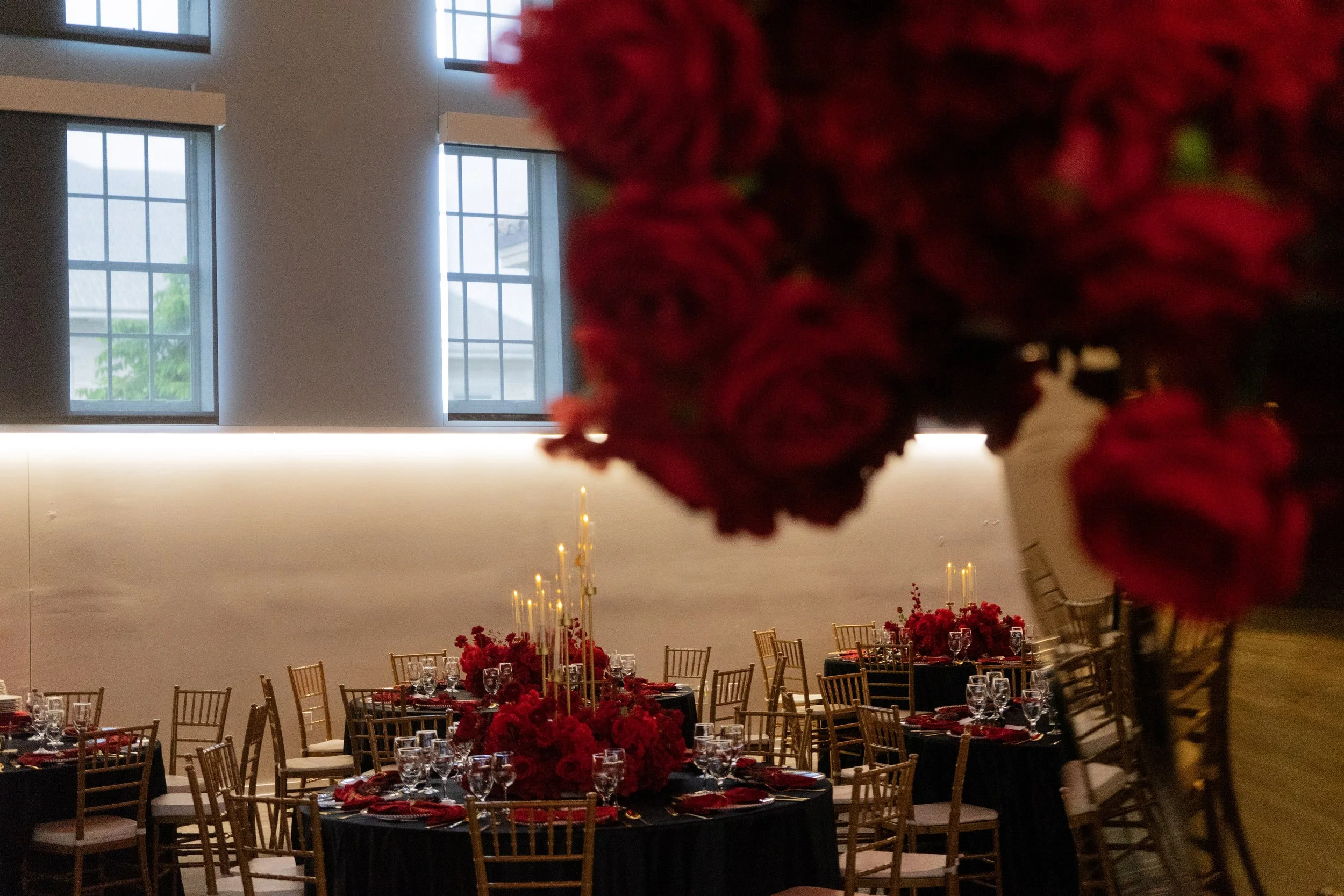 A banquet hall is decorated with round tables covered in black tablecloths, each adorned with red floral centerpieces and tall gold candles. Gold chairs surround the tables. Large windows with grid panes are visible on the wall, and soft lighting illuminates the room. In the foreground, part of a red flower arrangement is visible but out of focus.
