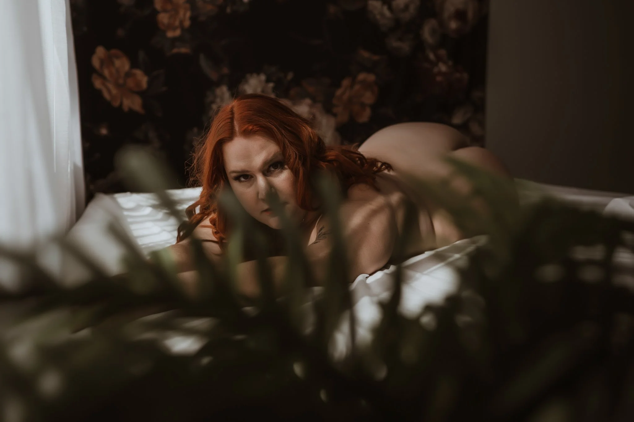 A red-haired woman lies on her stomach on a bed with white sheets, gazing directly at the camera with an intense expression. The scene is dimly lit, with a floral patterned headboard and dark wallpaper in the background. Green foliage is blurred in t
