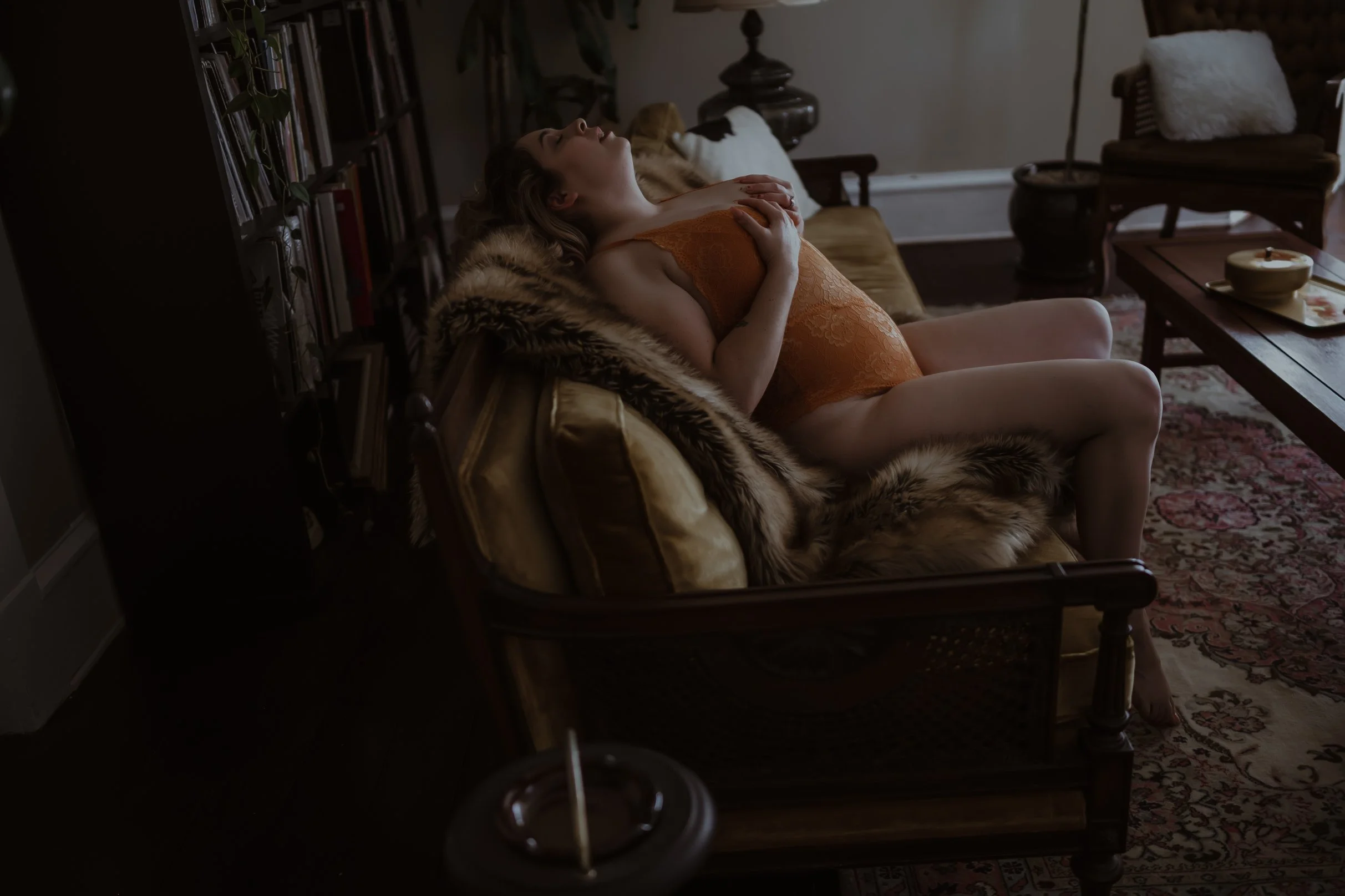 A woman with curly hair lying on a vintage-style couch with a fur throw, wearing an orange lace lingerie, in a dimly lit living room with wooden furniture and patterned rug.