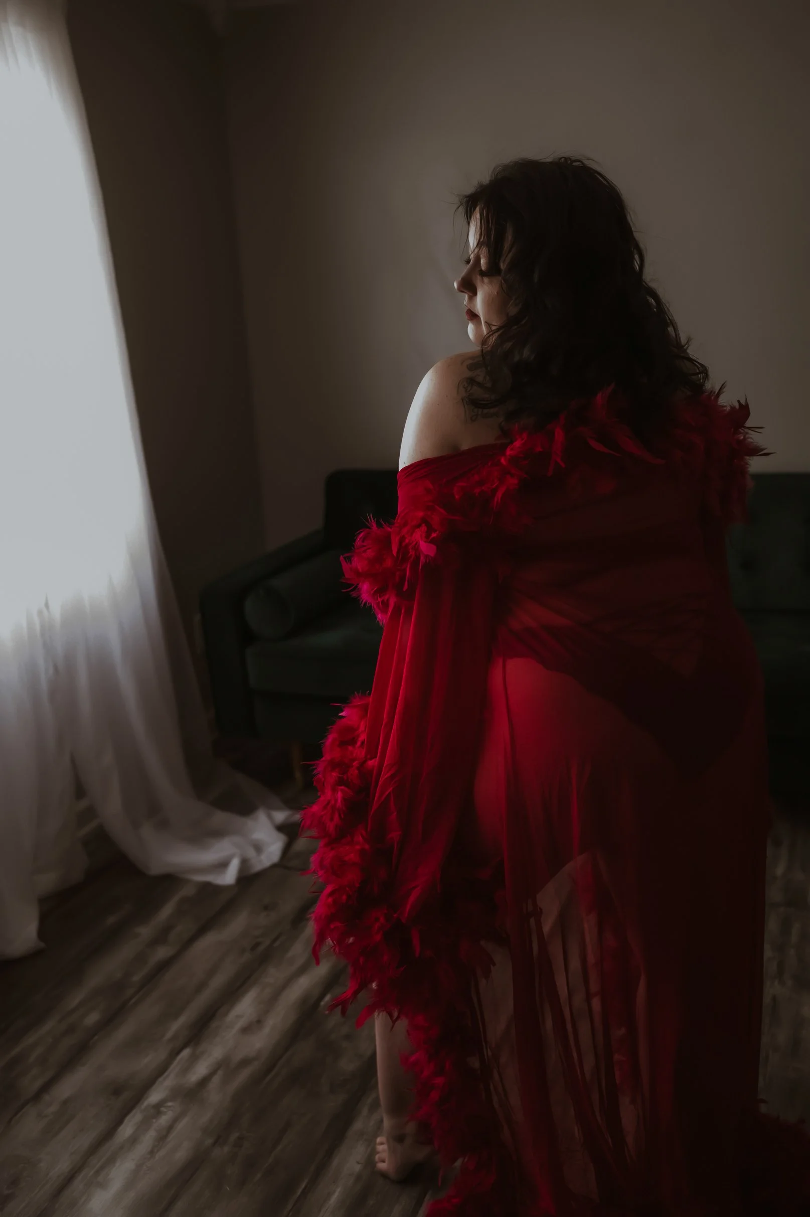 A woman with dark curly hair standing in a dimly lit room, wearing a red sheer robe with feathered embellishments, facing a window with sheer white curtains.