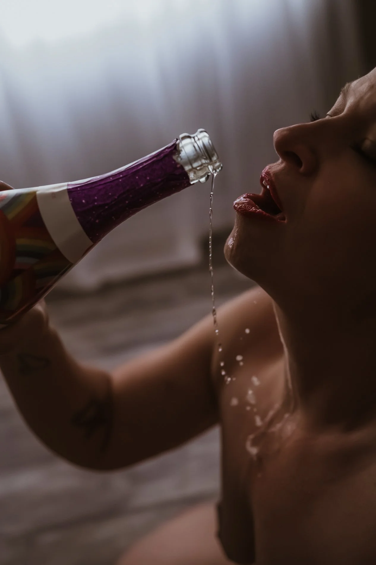Person with tattoos holding a bottle of purple liquid, pouring it into their open mouth.