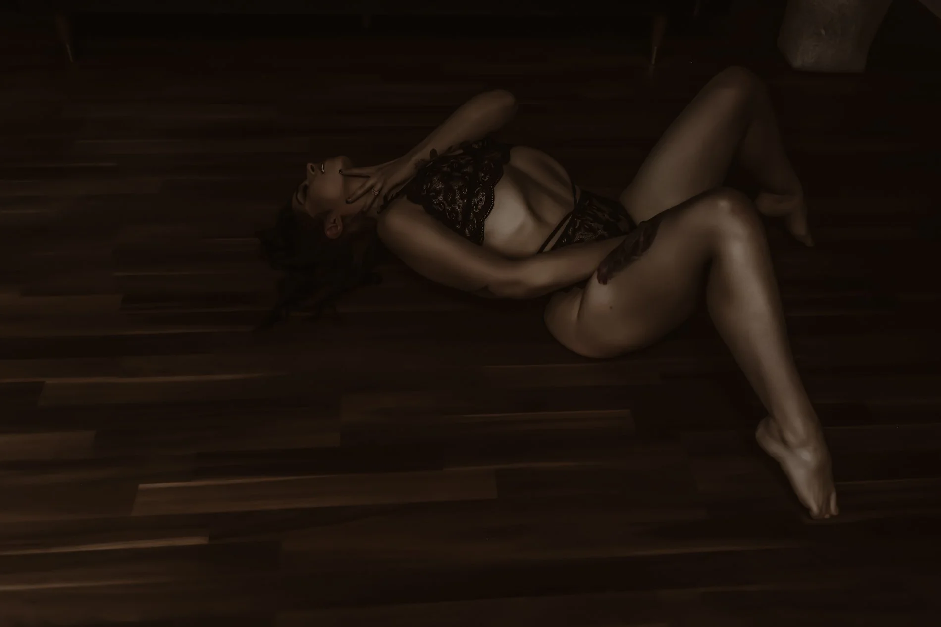 A woman lying on a wooden floor, wearing black lace lingerie, poses with one hand near her face and the other on her neck. The lighting is dim and atmospheric.