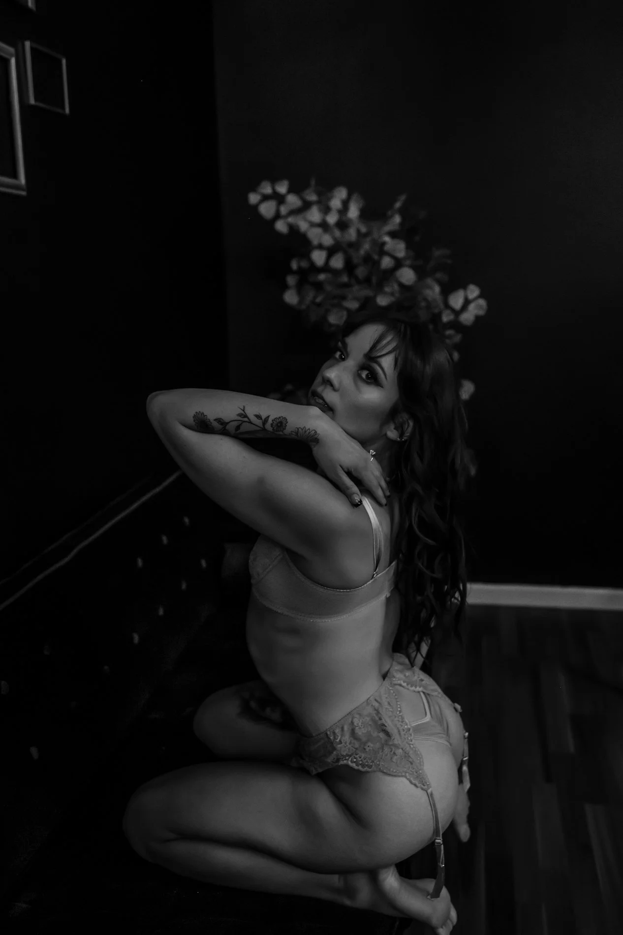 A woman with long, wavy hair poses on her knees with one arm across her face, looking at the camera in a dimly lit room. She is wearing lingerie, displaying tattoos on her arm and thigh, with a decorative floral wall behind her.