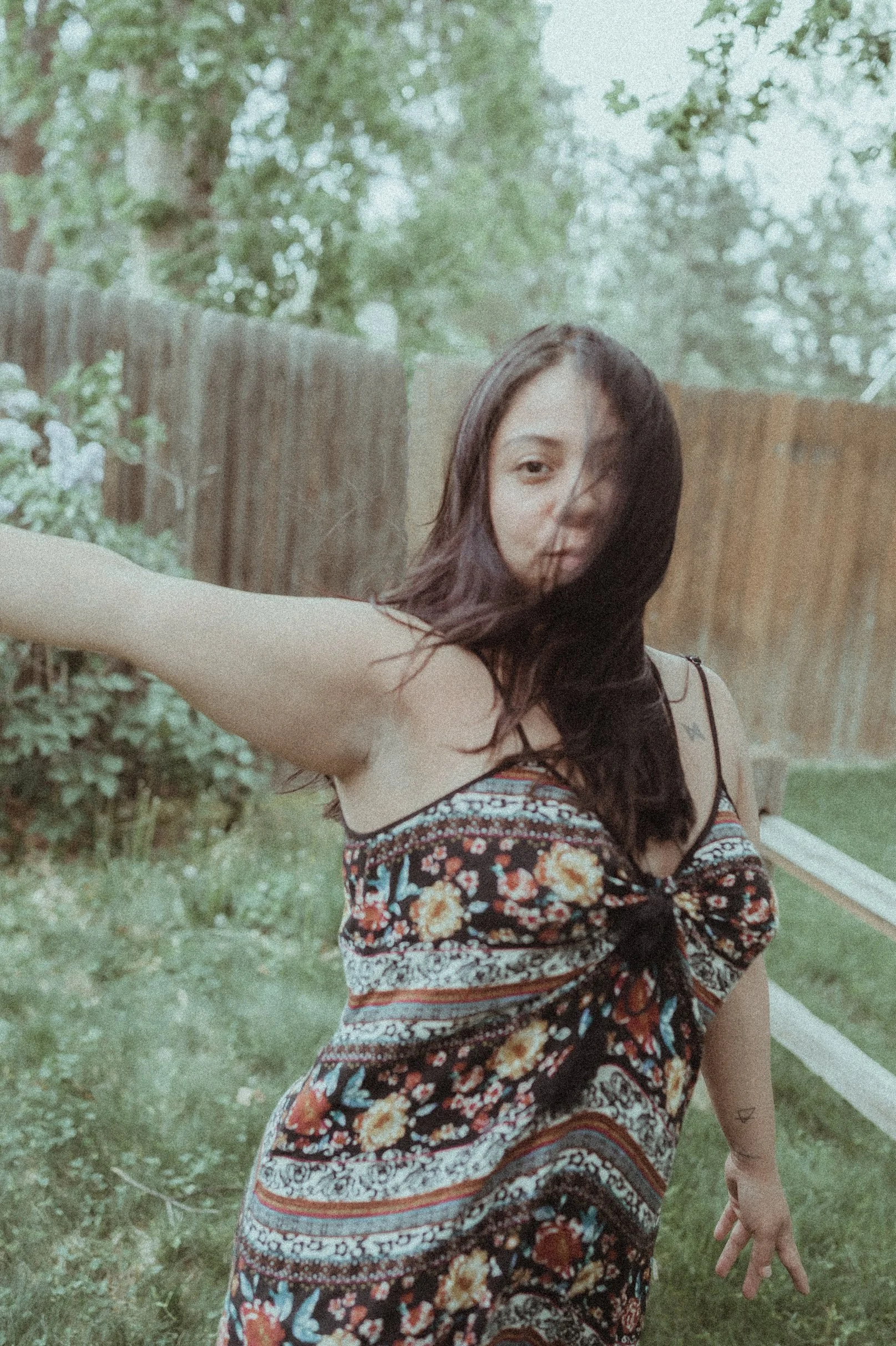 A young woman with long dark hair wearing a colorful floral dress taking a selfie outdoors in a backyard with green grass, bushes, and a wooden fence.