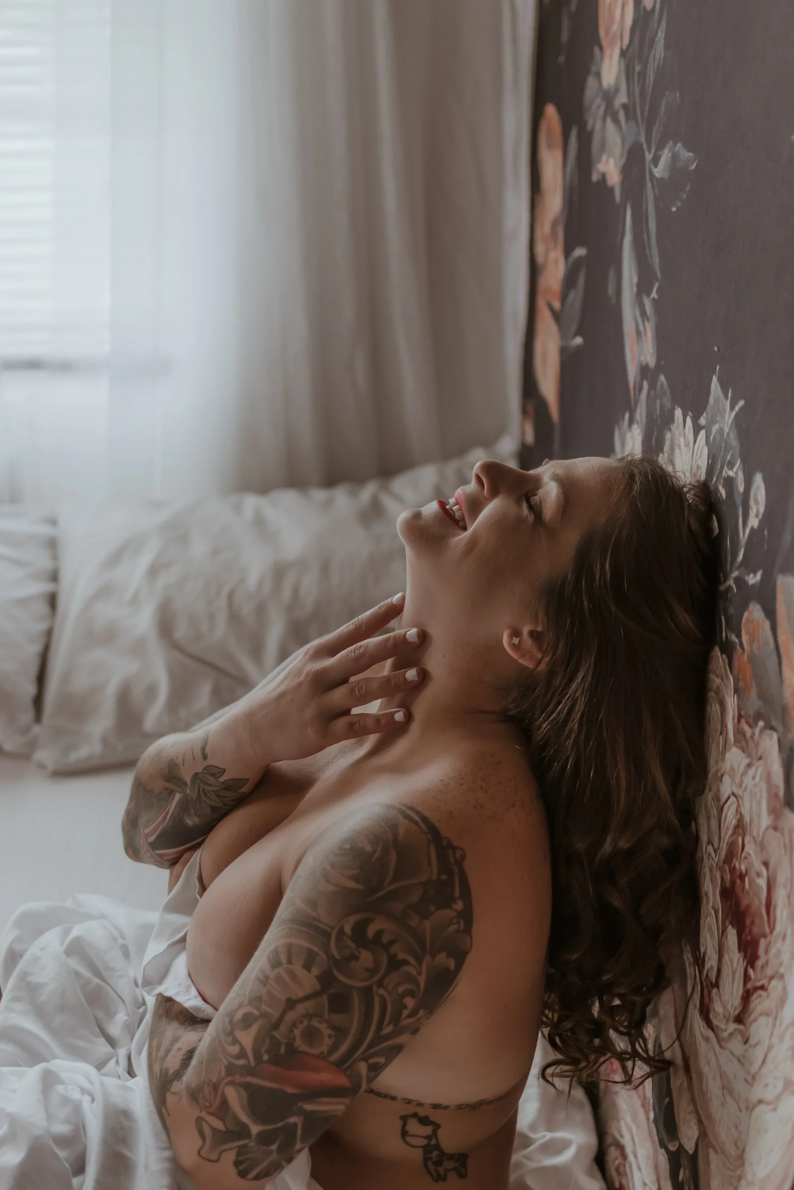 A woman with tattoos on her arms and shoulder, smiling and relaxing on her bed with her hand on her neck, leaning against a floral-patterned headboard in a softly lit room.
